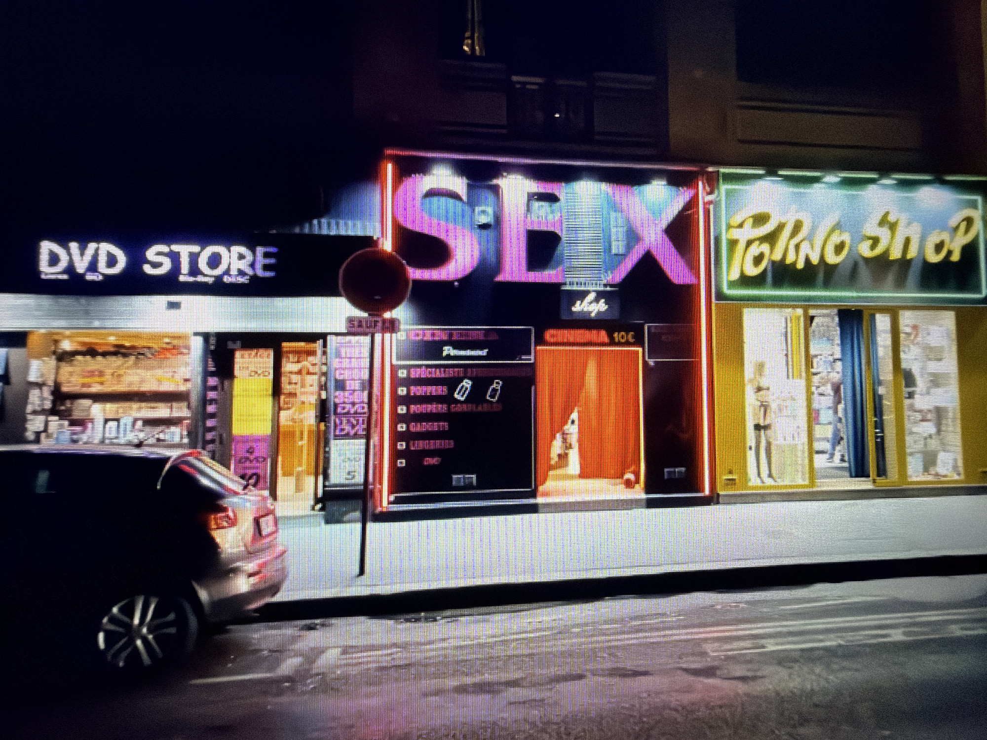 The image captures a street scene in an urban area at night. On the left is a "DVD STORE". Adjacent to it is a "SEX shop" with a neon sign. The store displays a list of products, including poppers and gadgets. To the right of the sex shop is a "Porno Shop". A car is parked in front of the shops. A traffic sign is present.