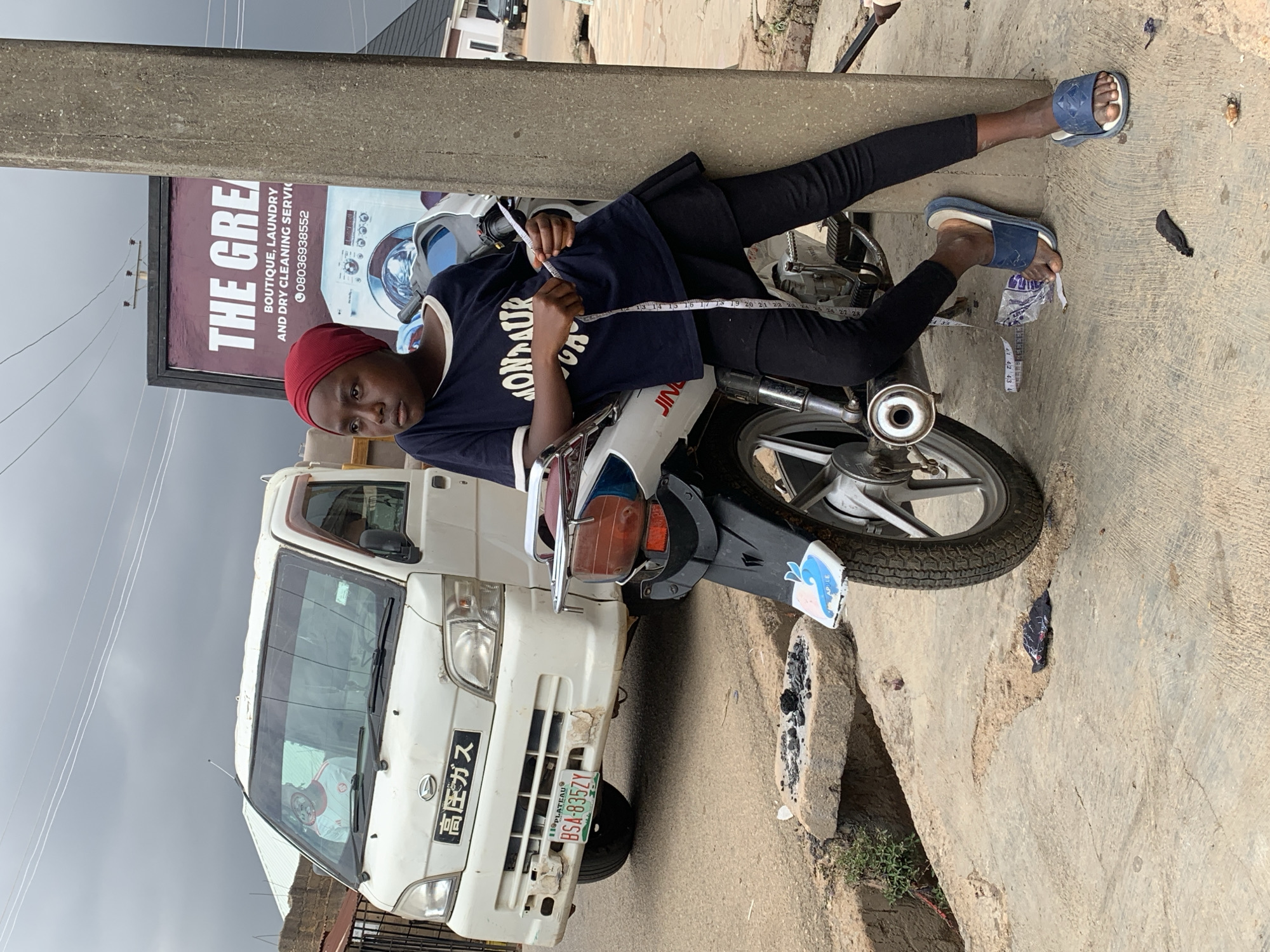 A person with a red head covering is sitting on a white motorcycle next to a white truck on the side of a road. The person wears a dark shirt with white writing and black pants. A measuring tape is wrapped around their body, presumably for measurements. They are wearing blue sandals. The motorcycle has a license plate on the back. A white truck is parked in front of the motorcycle, with a license plate and Japanese characters visible on the front. Behind the motorcycle and truck, there is a sign for "THE GREAT" laundry service. The location is Gura Top, Nigeria. The road appears to be paved but worn, with debris and uneven surfaces. A concrete pillar is nearby.