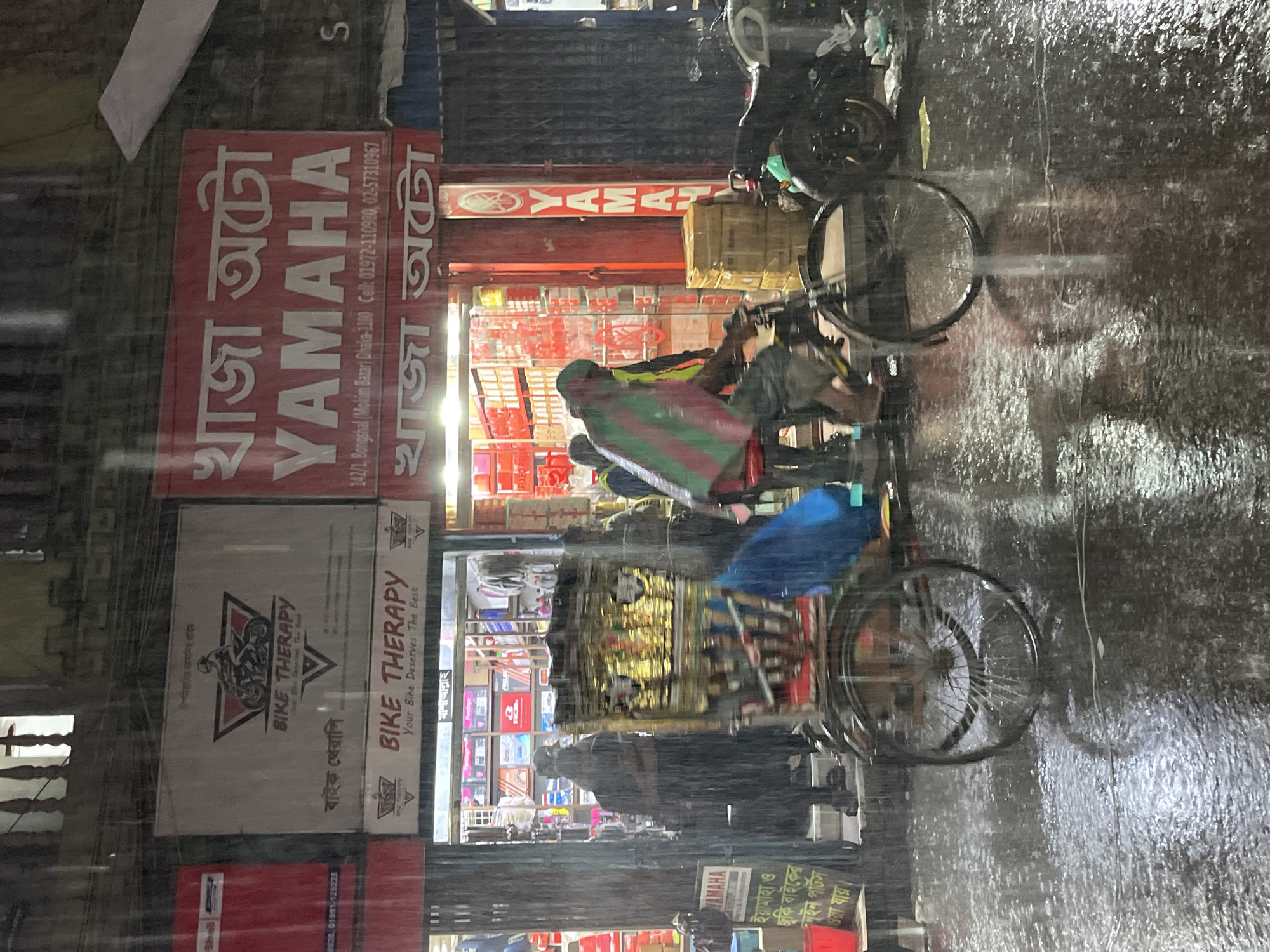 The image depicts a street scene in Dhaka, Bangladesh, during heavy rain. In the foreground, raindrops streak the view. The left side of the image features storefronts. One sign reads "BIKE THERAPY" and displays a motorcycle graphic. Above it, another sign reads "খাজা অটো YAMAHA" in Bengali and English, indicating a Yamaha dealership. The sign also provides the address "142/1, Bongshal (Molim Bazar) Dhaka-1100" and contact numbers. A person wearing a green and red striped covering is visible near the Yamaha store, and bicycles are parked along the side of the street. The reflections on the wet pavement add to the rainy atmosphere. The overall impression is one of a busy urban street scene affected by heavy rain.