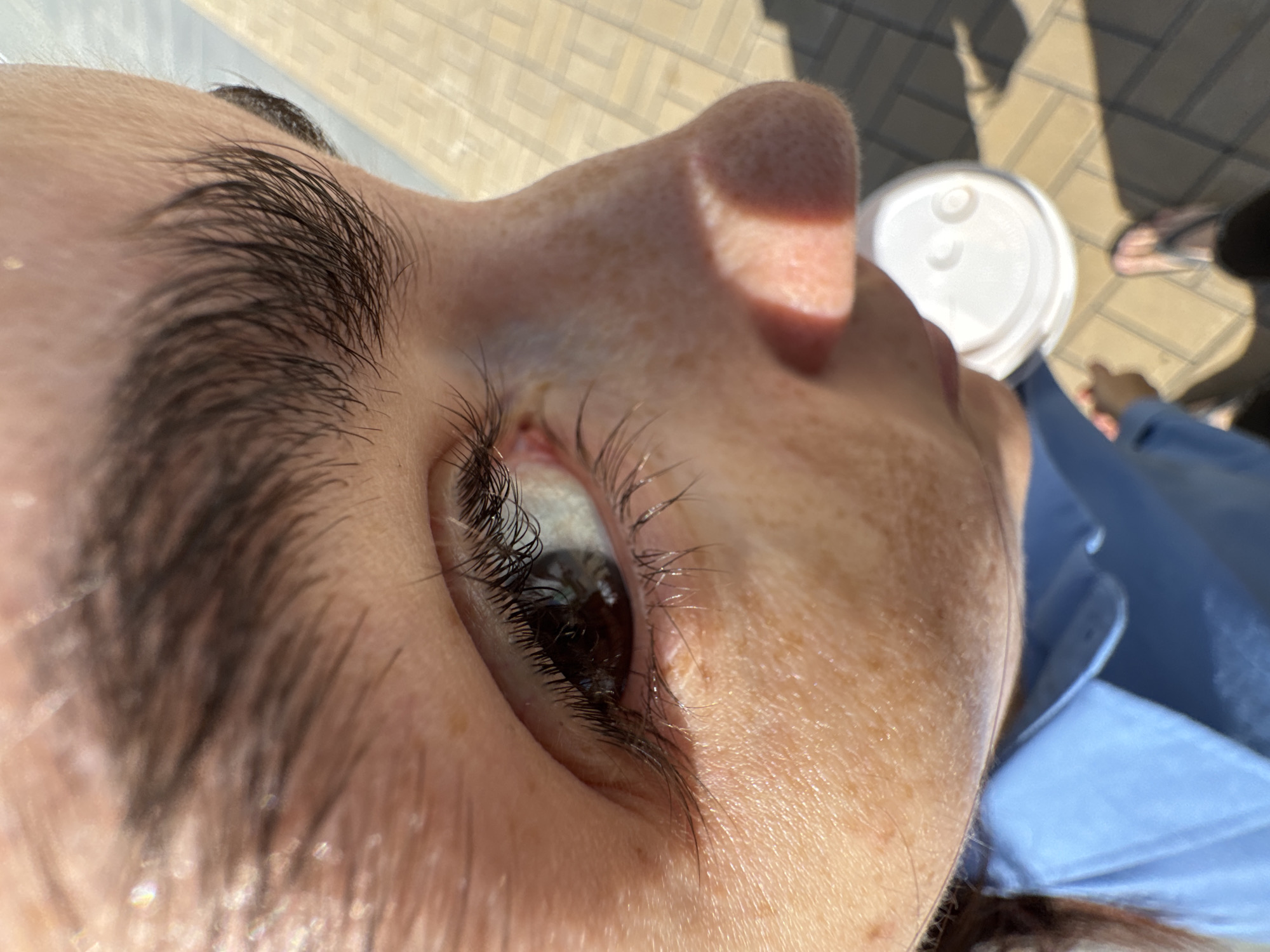 This extreme close-up, low-angle shot, taken in Tyumen, Russia, captures a person's face looking upwards under bright sunlight. The prominent features are the individual's right eye, eyebrow, and nose, visible from a worm's-eye perspective. The eye is open, showing a dark iris and long, dark eyelashes. A thick, dark eyebrow frames the eye, and the surrounding skin is fair with visible freckles. Direct sunlight casts distinct shadows across the nose and cheek.
In the blurred background, paving stones or tiles indicate an outdoor setting. A white plastic lid, likely from a cup or container, is visible in the middle-right. Further back, vague shapes suggest another person's feet or shoes. Part of a blue collared shirt is visible on the lower right. The scene is illuminated by bright daylight, implying sunny weather. The person appears to be relaxed, perhaps looking at the sky or something above them. No discernible text is visible on any objects in the image.
