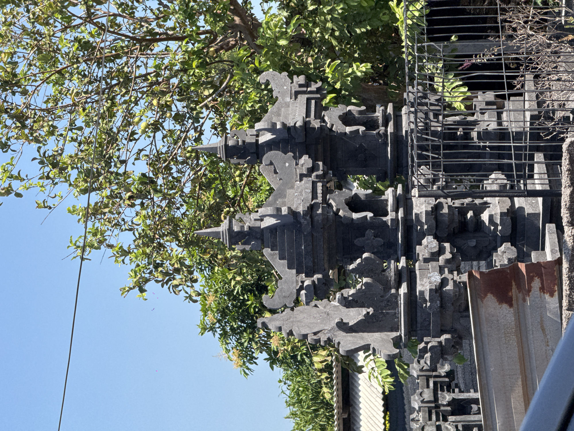The image shows an outdoor scene with a clear blue sky. In the foreground, there is a dark grey, intricately carved stone structure, possibly part of a temple or gate. Behind this structure, a lush green tree with some visible fruit or buds is prominent. To the left of the stone structure, a white corrugated roof is partially visible. A black metal fence runs across the bottom right of the image, partially obscuring some of the stone work and greenery behind it. The overall setting suggests a tropical or sub-tropical environment. Based on the architectural style of the stone carvings, it is likely located in Denpasar, Indonesia, a city known for its Hindu temples and traditional Balinese architecture.