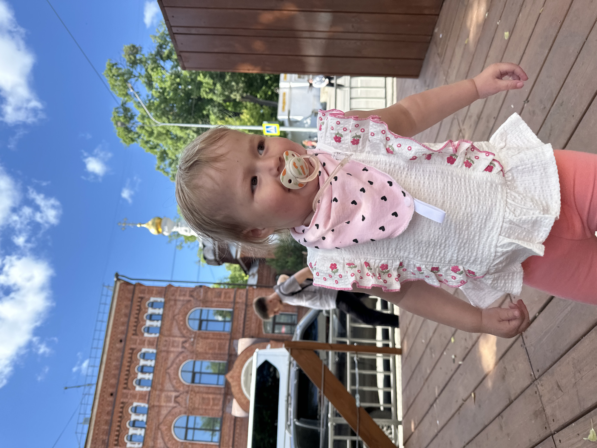 A brightly lit, outdoor photograph features a young toddler standing upright on a wooden surface, looking towards the camera. The child has fair hair and a pacifier with an orange-colored handle in their mouth. They are dressed in a white smocked top with ruffled sleeves and pink floral patterns, a pink bib decorated with small black hearts, and light pink or coral trousers. Their right arm is slightly raised.
In the background, under a clear blue sky with scattered white clouds, a red brick building with arched windows is prominent. Above and to the left of this building, a golden church dome topped with an Orthodox cross can be seen. Green trees are also visible. A partially obscured adult figure is visible further back, near what appears to be a vehicle and some metal railings. The setting is a sunny day in an urban or historical area, consistent with the provided location context of Nizhny Novgorod, Russia. A yellow street sign is faintly visible in the mid-ground, but any text on it is unreadable.