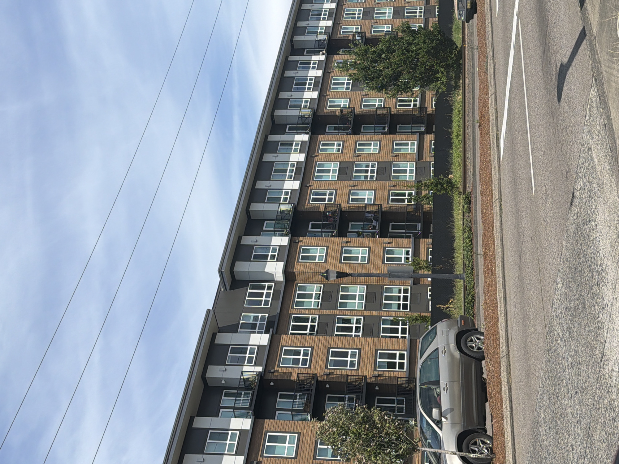 An exterior, eye-level, medium shot captures a modern, multi-story apartment building in Beaverton, Oregon, under a partly cloudy sky. The building features a dark gray roof and upper facade, with the majority of its exterior clad in horizontal, light brown wood-like siding. White-framed windows are arranged in a grid pattern across the facade, with some windows on the upper floors featuring small, black-railed balconies. Several trees, including a mature evergreen and a smaller deciduous tree, are situated in front of the building, interspersed with landscaping. A light-colored SUV is parked in front of the building on the left side of the frame. The foreground shows a paved street with white lane markings. Power lines are visible against the sky.