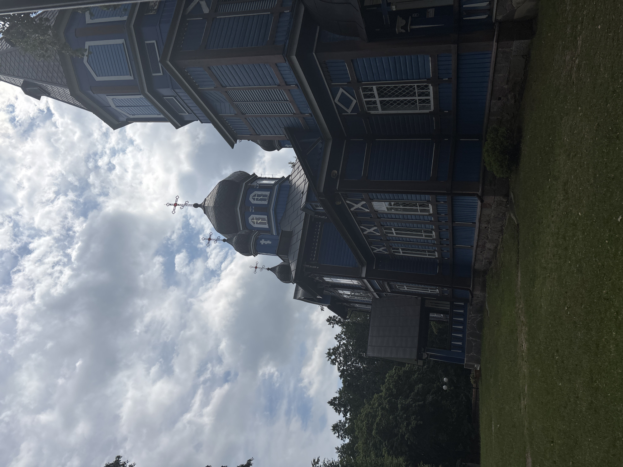 The image shows an exterior view of the Orthodox Church of the Nativity of the Blessed Virgin Mary in Puchły, Poland. The church is made of wood and is painted blue with dark brown trim. It has multiple onion domes topped with crosses. The church is situated on a grassy area with trees in the background. The sky is cloudy. The perspective is from the side of the church, looking up towards the domes and spire. The building's architecture features decorative window frames and horizontal wooden siding. The base of the church appears to be made of stone. There is a porch or entryway visible on the left side of the building.