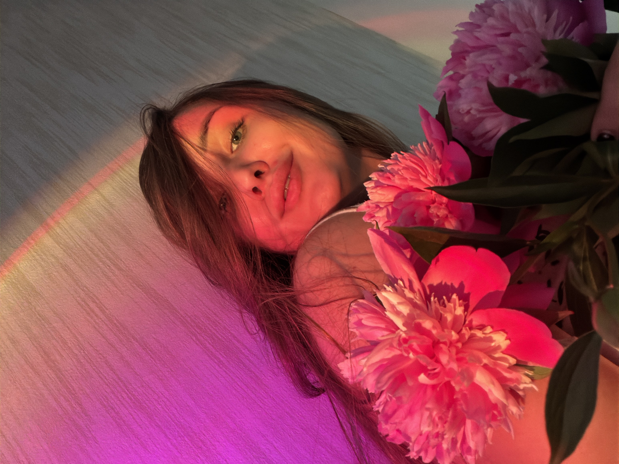 A young woman with long, dark hair and green eyes is captured in a close-up shot, holding a bouquet of pink peonies. She is looking towards the camera with a slight smile, her face illuminated by a pinkish-orange light that casts a gradient across the background. A faint rainbow-like light streak is visible in the upper right portion of the image. The background appears to be a textured wall. The scene is lit with warm, vibrant colors, suggesting it might be sunset or dawn. There are no identifiable landmarks or contextual clues to pinpoint the location within the city of Chaikovsky, Russia. The image is a portrait with a focus on the woman and the flowers.
