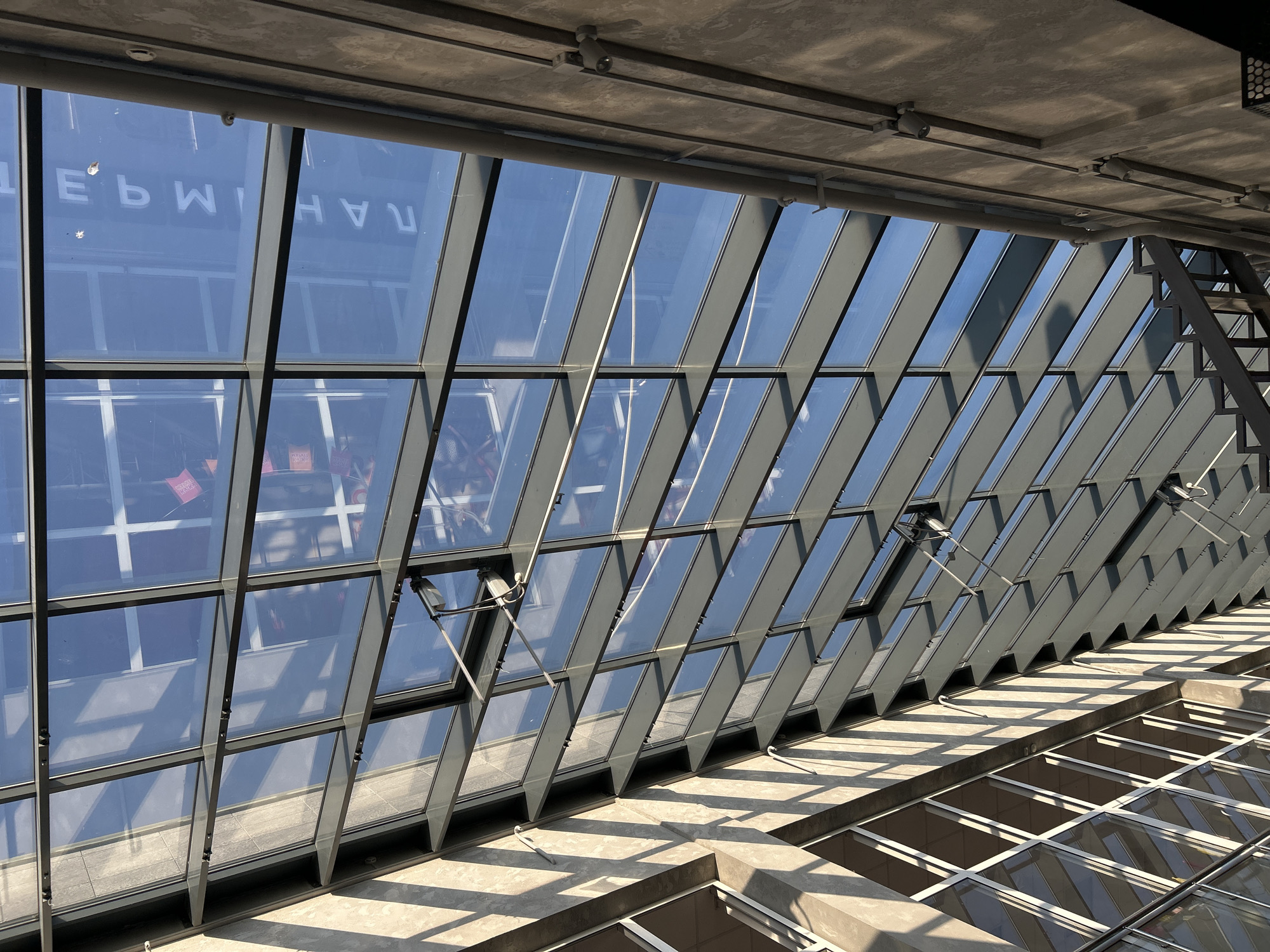 The image shows a view looking up at a modern glass and steel atrium ceiling. Sunlight streams through the glass panels, casting long, angled shadows onto a concrete wall and the structural elements of the ceiling. The reflection in the glass shows a blue sky and some faint white lettering, possibly advertising or signage, which reads "ТЕРМИНАЛ" (TERMINAL). This suggests the location is a transportation hub or a large commercial building. Based on the text, the image is located in Izhevsk, Russia. There are no people clearly visible in the image, but there are some shadowy shapes reflected in the glass that could indicate activity within the building. The overall impression is one of modern architecture and the interplay of light and shadow.