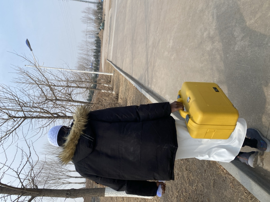 A single individual is observed from behind, walking on a paved surface in Zhongtun, China, under a clear sky. The person is wearing a white kufi cap, a black hooded winter jacket with a brown fur-trimmed hood, a white long garment extending below the jacket, dark lower garments, and dark athletic shoes. In their right hand, they carry a rectangular, bright yellow hard-shell equipment case with a grey handle. In their left hand, they hold a rolled-up white or light blue object, possibly a document or blueprint. To the immediate left of the paved path is a raised dirt embankment with several bare deciduous trees. Further in the background, more bare trees line a distant perimeter. A white street lamp post with a blue-grey light fixture is visible on the right side of the path. In the far distance, industrial structures and power lines are discernible against the horizon. The ground consists of grey asphalt or concrete, with dry earth and sparse vegetation on the embankment.