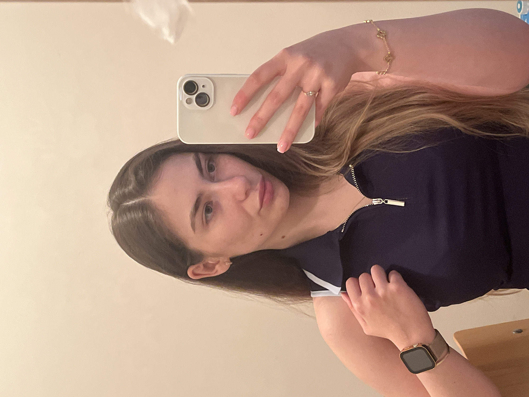 A mirror selfie captures a young woman from the chest up, reflected in a mirror. Her body is oriented vertically, head to the left, as she looks towards the viewer. She has long, dark brown hair with lighter undertones, styled with a side part, flowing over her shoulders. Her face is turned slightly to the right, revealing light-colored eyes and a neutral, calm expression.
She holds a light-colored smartphone, possibly an iPhone, with her right hand in front of her face, its camera lenses visible on the back. Her right hand also features a delicate gold-colored ring with a small stone on her ring finger, and her fingernails have a light pink ombre manicure. On her left wrist, she wears a gold-colored bracelet adorned with small, possibly clover-shaped charms, and a square-faced smartwatch with a gold frame and a dark band.
The woman is dressed in a dark blue top, likely a polo shirt, with a visible white collar and white trim on the sleeves. A metallic zipper with a rectangular pull tab is prominent at the neckline. The background is a plain, light beige or cream-colored wall, suggesting an indoor setting. A faint, white, irregular shape is visible in the upper left corner, possibly a reflection or light fixture. Part of a wooden surface, such as a table or shelf, is visible in the bottom right corner. No visible text is present in the image.