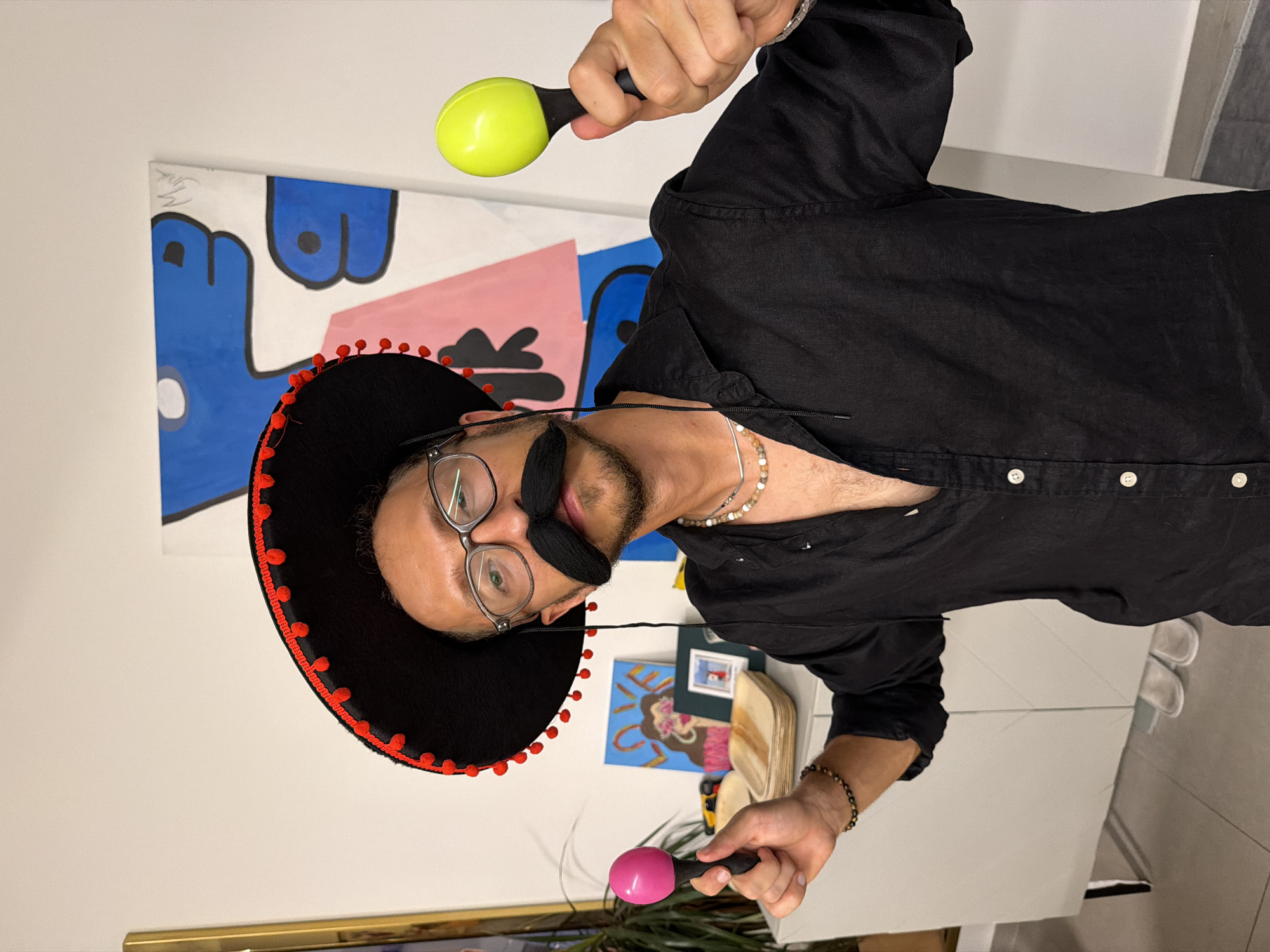 A man in his 30s, wearing a black sombrero with red pom-poms, glasses, a fake mustache, and a black button-down shirt, poses in front of a white wall with abstract art. He holds two maracas, one pink and one lime green, in his hands, as if playing them. In the background, on a white dresser, are several items including a framed picture, a plant, and stacks of square, light-brown plates. The man's left arm is slightly raised, and his right arm is extended forward, holding the green maraca. The lighting suggests an indoor setting, possibly a home or studio. There are no indications of Tel-Aviv, Israel in the image.