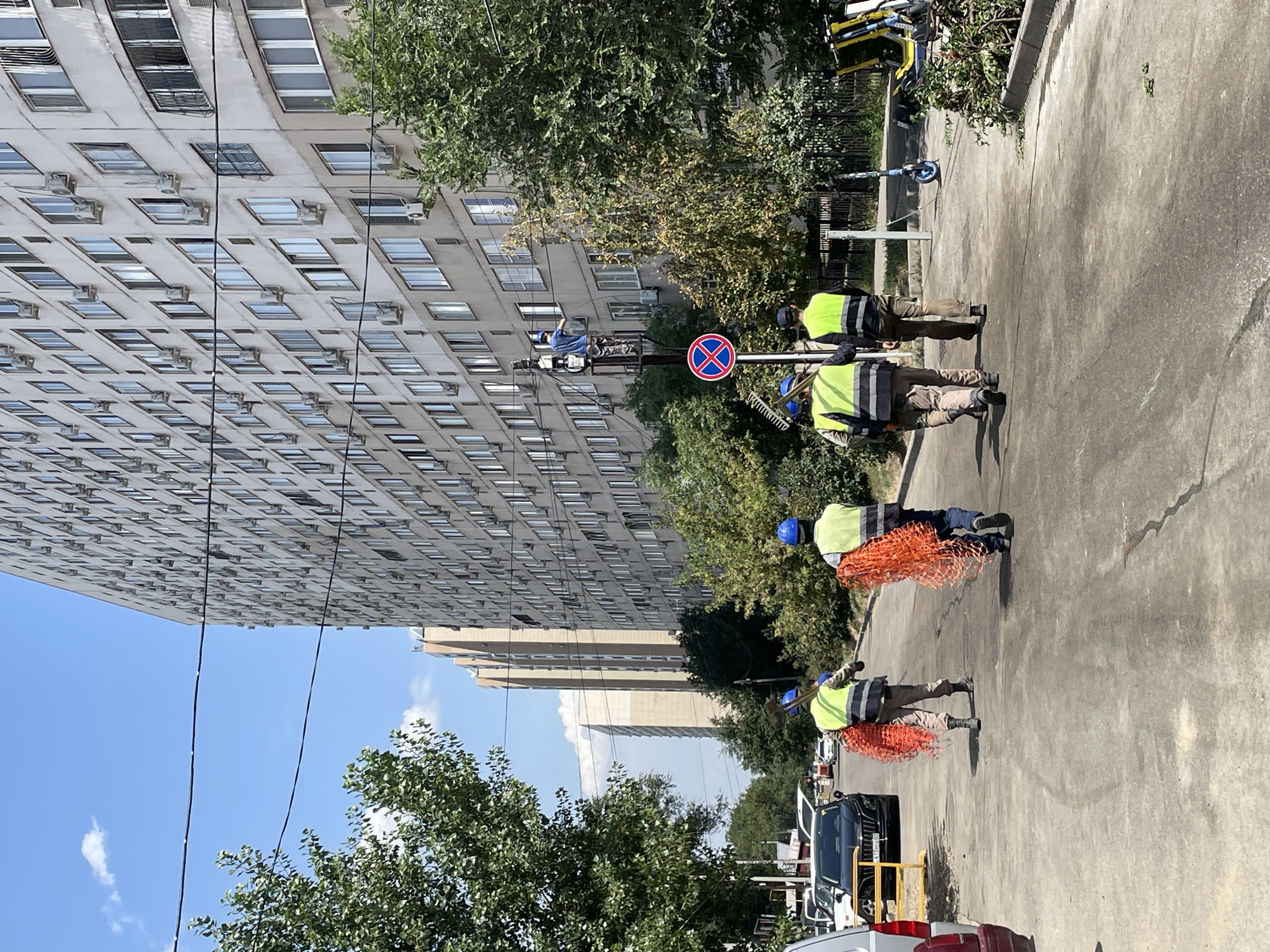 In Almaty, Kazakhstan, a group of workers in reflective vests and hard hats are seen on a paved road in front of a large apartment building. Two workers in the foreground carry orange netting. Further down the road, another worker is on a utility pole, possibly performing maintenance or repairs, while two more workers stand at the base of the pole. A traffic sign with a blue circle and red cross is visible near the pole. The environment appears to be a residential area with trees and parked cars, including an electric scooter and a yellow electric bike in the background. The sky is blue with scattered clouds.