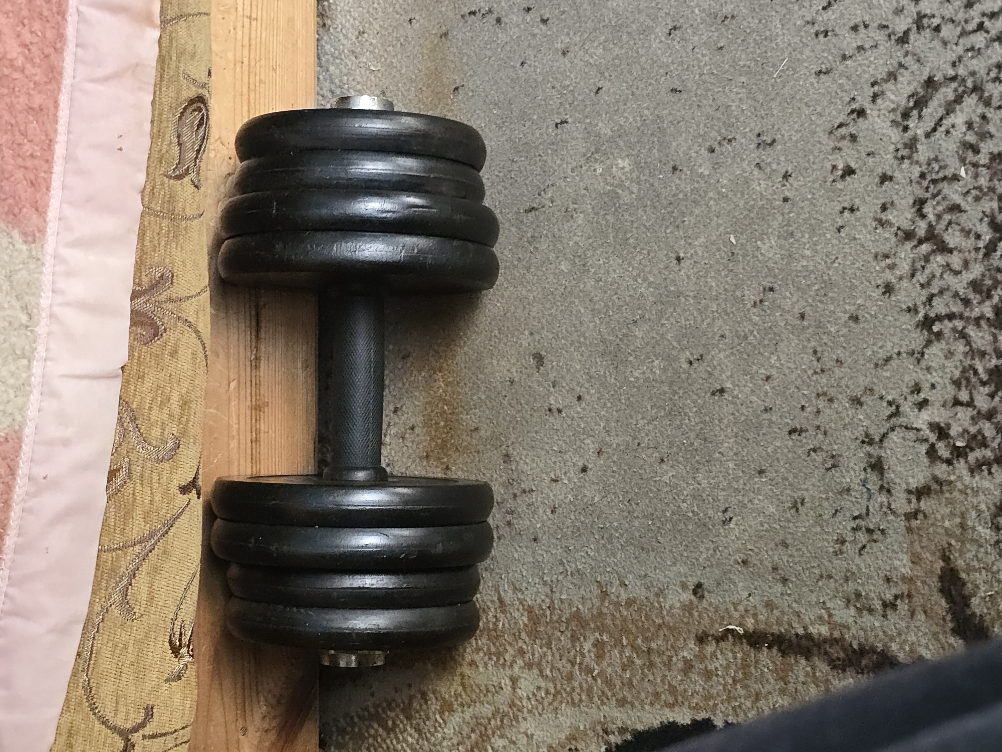 A dumbbell is resting horizontally on a piece of wood, which is itself lying on a carpeted floor. The dumbbell has black weights on either end of a textured black handle. The weights are made of multiple circular plates. Above the dumbbell and wood is a piece of fabric with a light pink trim and a patterned yellow fabric. The carpet is a mottled grey and brown, with some darker stains and specks. The scene appears to be indoors, in a room or perhaps a garage.