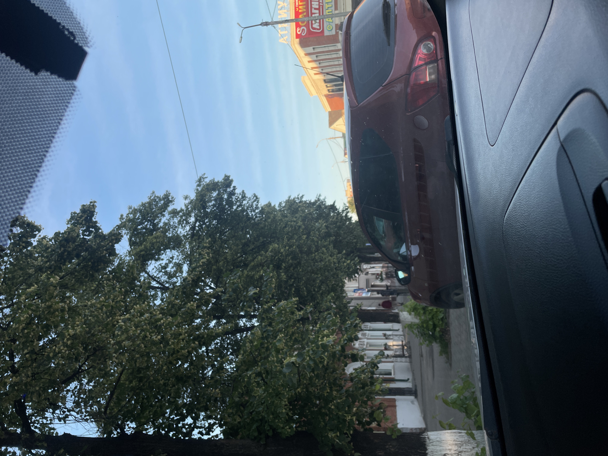 An outdoor, eye-level, slightly low-angle shot shows a street scene in Votkinsk, Russia, on a bright, cloudy day. The image is taken from inside a vehicle, looking out through the windshield.
In the foreground, the dark, textured dashboard of a car dominates the bottom of the frame. The windshield itself shows reflections of the sky and parts of the car's interior, including a black, triangular object and a patterned texture at the top.
The middle ground is occupied by a street with a sidewalk and trees lining the left side. The trees are full of green leaves and appear to be mature. The sidewalk has a paved surface and is partially obstructed by greenery. On the right side of the street, a maroon hatchback car is parked or in traffic. Its rear is visible, including the taillights and a reflective surface on the back.
Further down the street, buildings with red and white facades are visible. One building on the right has a prominent sign with Cyrillic lettering, partially obscured by an antenna. The letters appear to be "АТИ НУ" and "SCAL" followed by "МАГНІ" and "О АЛТІ". Streetlights are present along the street.
Two individuals are visible on the sidewalk in the middle distance. One person, wearing red, is walking away from the camera. Another person is also present but less distinct.
The background consists of a clear blue sky with faint wispy clouds. The overall impression is a typical urban street scene.