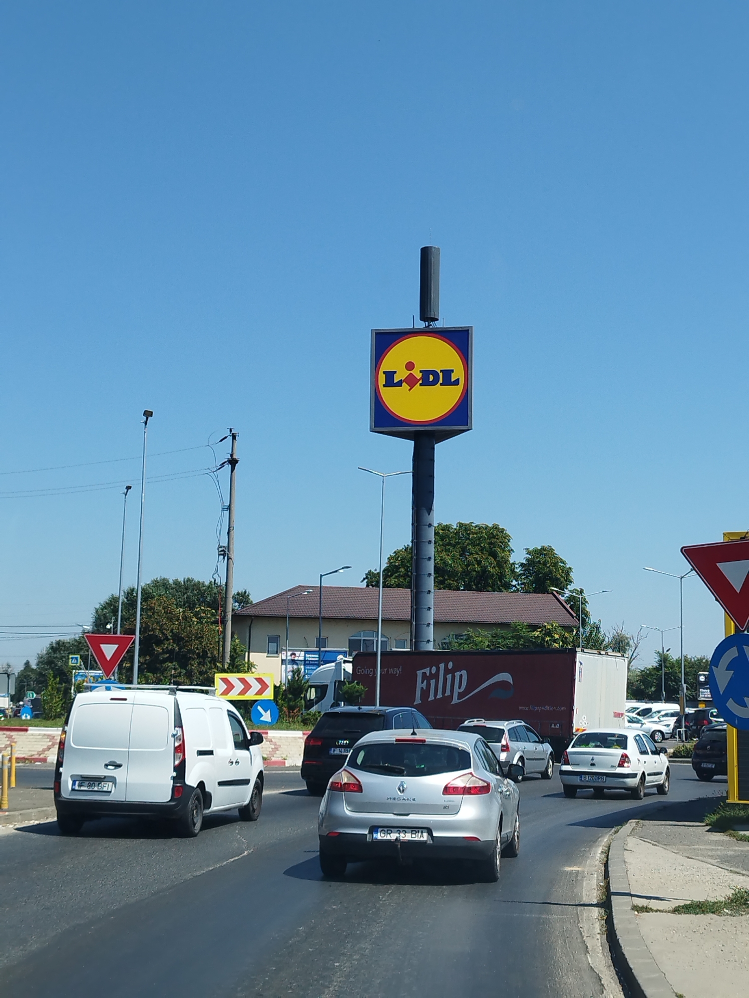 The image captures a street scene in Bragadiru, Romania, on a clear, sunny day. A large, square Lidl sign with its yellow and blue branding is prominently displayed on a tall pole, acting as a landmark. Below the sign, a red and white truck with "Filip" branding is visible. The road is busy with traffic, featuring a white van, several cars including a silver Renault Megane, and other vehicles. Various traffic signs are present, including a yield sign and a roundabout sign. The surrounding environment includes buildings with brown roofs, trees, and streetlights. The sky is a bright, cloudless blue.