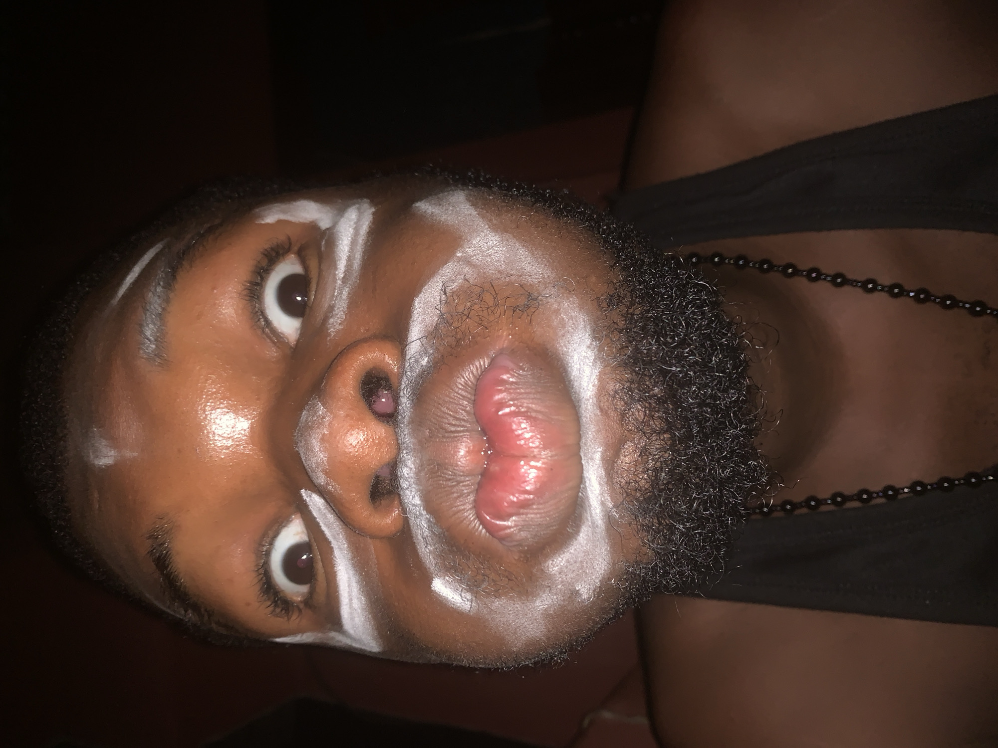 A close-up, rotated portrait shot features an individual with dark skin and a full, dark beard, against a dark background. White paint or chalk marks are applied in streaks across the forehead, around the eyes, down the nose, and along the cheekbones and chin, creating a distinct facial pattern, possibly for cultural expression or costume. The person's dark eyes are wide open and directed at the viewer, conveying an intense and alert gaze. Their lips are puckered tightly outwards, forming a prominent "kiss" or "fish" face, with the moist, red interior of the lips clearly visible. A dark, beaded necklace is visible around the person's neck, partially obscured by the beard. A black garment, likely a tank top strap, is seen on the right side of the frame, on the person's shoulder/chest area. The lighting is direct, illuminating the face and highlighting the texture of the skin, the white markings, and the beard. The dark background suggests the photo was taken at night or in a dimly lit indoor environment. No text is visible in the image. The provided context indicates the location is Onitsha, Nigeria.