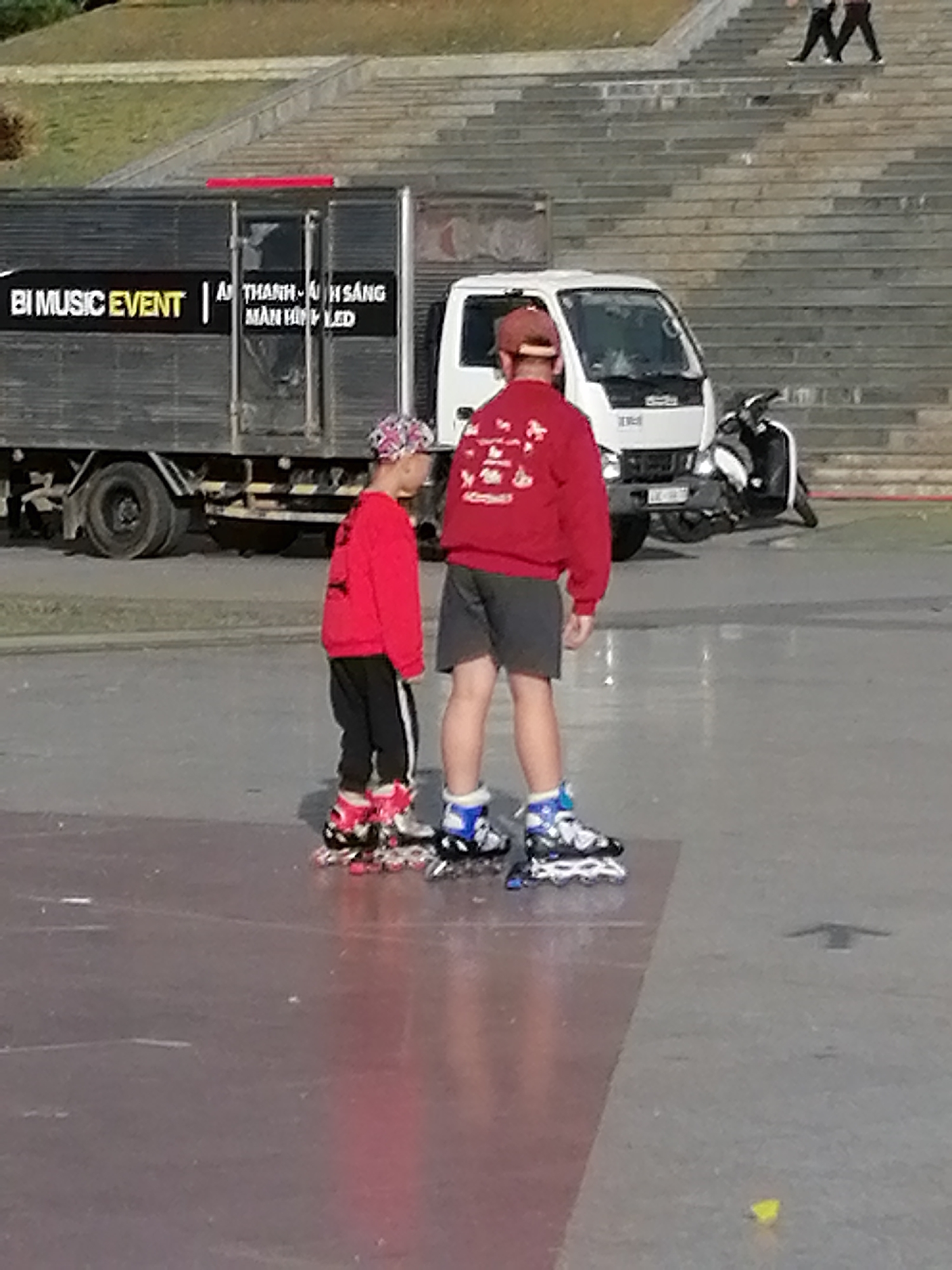 Two young people are inline skating on a paved area. The person on the left is wearing a red sweatshirt with a patterned hat, black pants with white stripes, and colorful inline skates. The person on the right is wearing a red sweatshirt, dark shorts, and blue and white inline skates.
Behind them, a large gray truck with "BI MUSIC EVENT" written on its side is parked. A white truck is also visible, as is a black and white scooter. In the background, a set of wide, stone stairs ascends a gentle slope. The scene appears to be outdoors, possibly in a public space. The lighting suggests it could be daytime, with the sun casting some reflections on the pavement.
The overall mood seems casual and recreational. There is no visible text related to time of day, weather, or specific emotions. The location cues like the stairs and the type of vehicles suggest an urban or park-like setting in Da Lat, Vietnam.