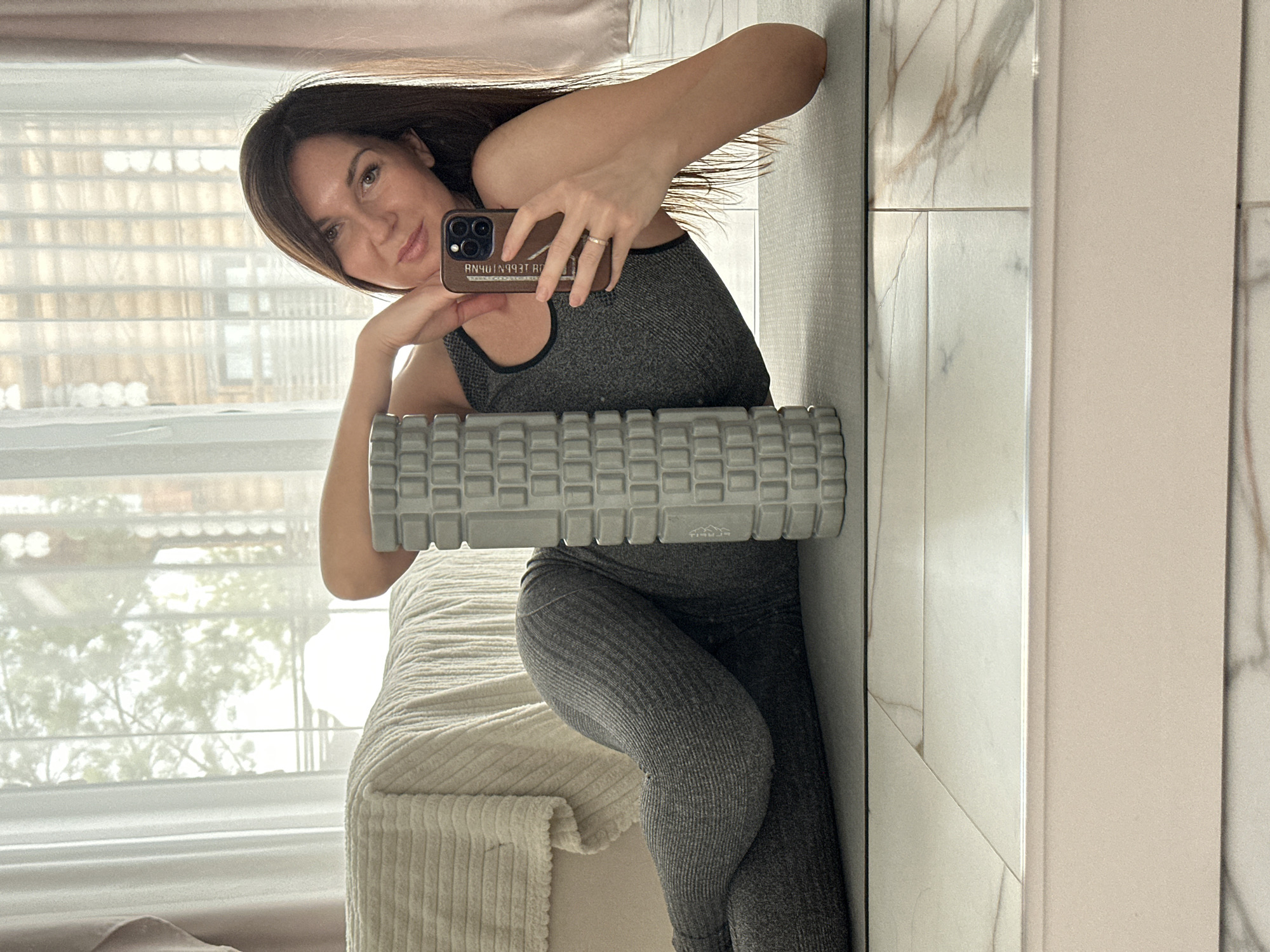 A female is depicted in an indoor setting, observed via a mirror reflection. She is positioned on her left side on a grey yoga mat, partially supported by her left elbow. She wears a grey ribbed sports bra and matching ribbed leggings. Her right hand holds a dark-colored smartphone with a brown case, angled towards herself. Reversed Russian text is discernible on the phone's screen or case. A light-colored ring is present on her left ring finger. A grey, textured foam roller stands vertically on the yoga mat, positioned between her torso and her bent right leg. The background features a large window covered by white sheer curtains, through which blurred trees and structural elements of a building are visible. To the left, a bed or couch draped with a light beige, textured blanket is present. The floor is covered with light-colored tiles exhibiting grey and brown veining. The scene is illuminated by natural light. The specified location is the city of Chaikovsky, Russia.