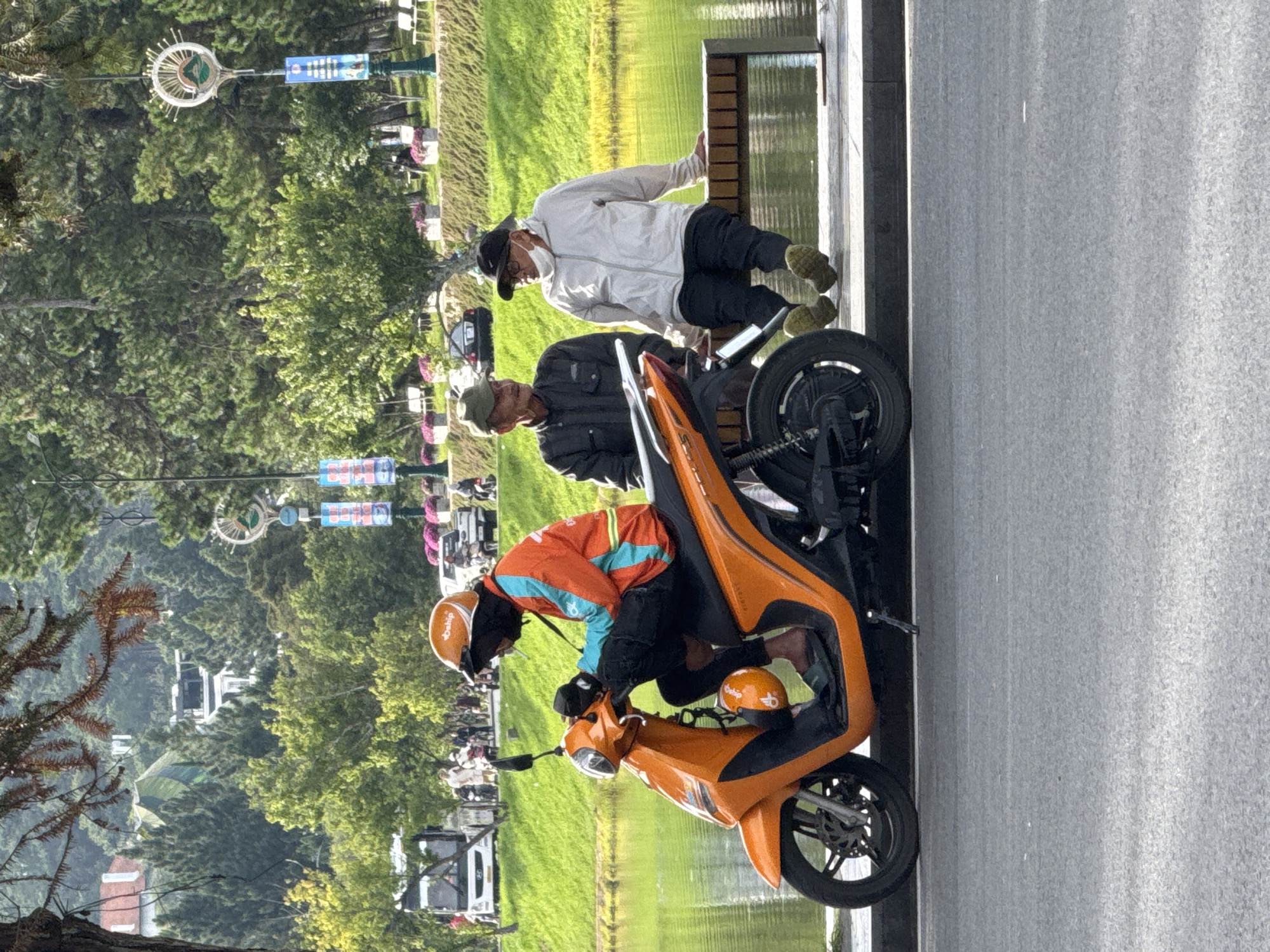 The image depicts a scene in Dalat, Vietnam, likely near Xuan Huong Lake given the presence of the lake and the surrounding park-like setting with trees and lamp posts. In the foreground, a person wearing an orange and teal jacket and an orange helmet is seated on an orange scooter. They appear to be smoking. To the right, two men are seated on a wooden bench by the lake. The man on the left wears a dark jacket and a green cap, while the man on the right wears a light-colored jacket, a black cap, and a face mask. The scooter is parked on the asphalt road, with the lake and a grassy slope with trees in the background. Various vehicles and pedestrians are visible in the distance.