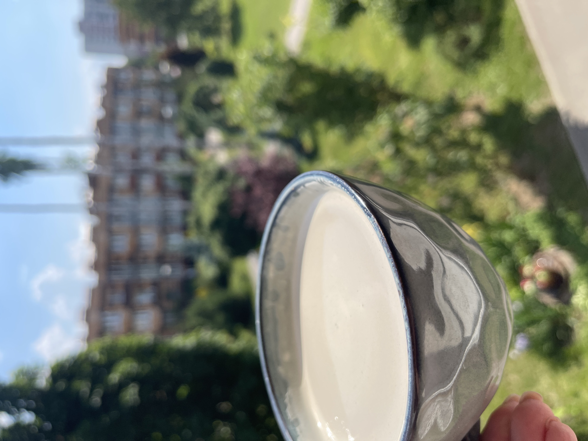 A close-up shot features a hand holding a grey, textured ceramic bowl filled with a pale, creamy liquid, possibly yogurt or a smoothie. The bowl has a dark blue rim. In the background, a blurred urban landscape is visible under a bright, sunny sky with scattered clouds. Several multi-story buildings, some with visible windows and balconies, are present. Lush green trees and manicured park areas with pathways indicate an outdoor setting. The overall scene suggests a moment of enjoyment of a refreshing drink outdoors, with a view of a residential area and park. It is not possible to determine the specific location of Kommunarka, Russia from the image alone.