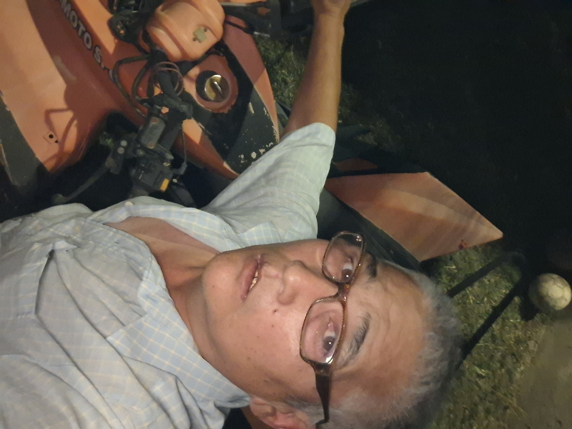 In a dimly lit outdoor setting, a middle-aged East Asian male with graying hair and glasses is pictured. He is wearing a light blue, checkered collared shirt. He appears to be sitting on or near an orange all-terrain vehicle (ATV). Part of the ATV's body, handlebars, and what appears to be a gas cap are visible. A weathered, off-white ball rests on a dark surface in the background, along with some green foliage. The overall scene suggests a casual, possibly domestic, environment in Almaty, Kazakhstan, though no specific landmarks are identifiable. The lighting indicates it is nighttime.