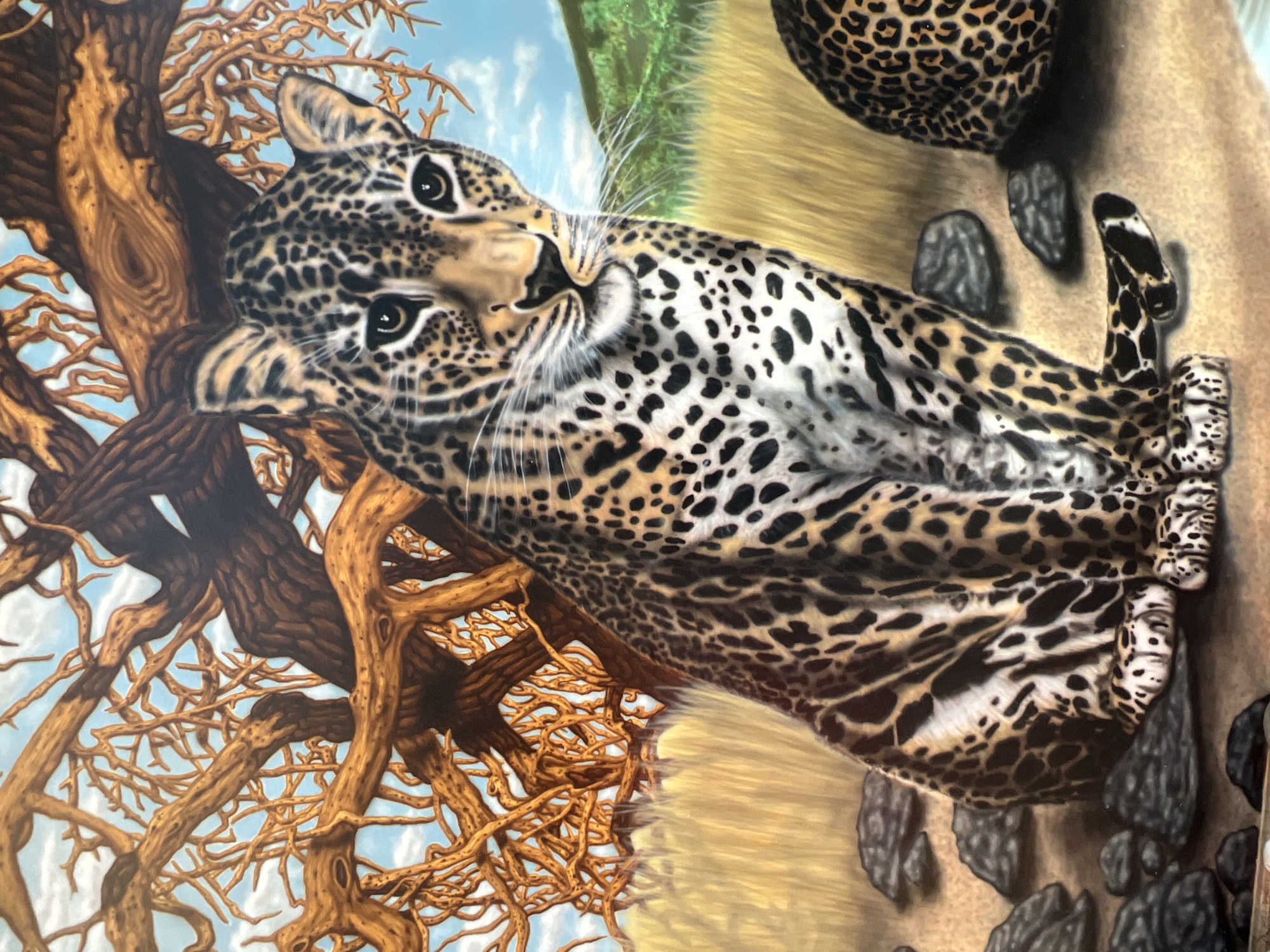 This is a painting of a leopard sitting on the ground in a savanna-like environment. The leopard is in the foreground, facing the viewer and looking slightly to the right. It has characteristic black spots on its tan fur. Behind the leopard, there is a gnarled, leafless tree with brown branches and a blue sky with white clouds. The ground is sandy with scattered rocks, and dry grass is visible in the background. Another leopard's hindquarters are partially visible on the right side of the frame. There is no discernible location in the city of Bender, Moldova depicted in this image.