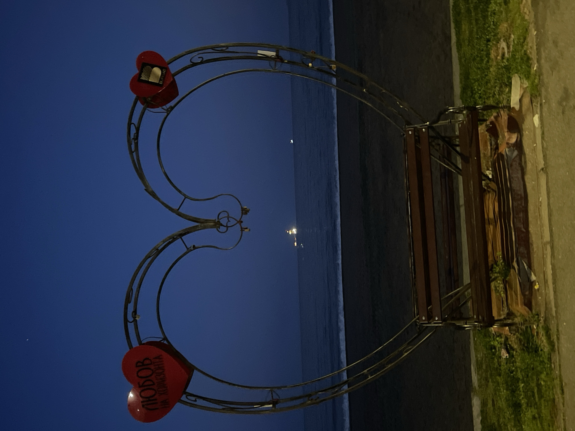 A night scene shows a heart-shaped wrought iron structure with a wooden bench, positioned on a paved area adjacent to a dark sandy beach and the sea. The sky transitions from a dark blue to black. Two red heart-shaped signs are attached to the top of the heart, one with the word "Burgas" and the other with the inscription "ЛЮБОВ на хоризонта" (Love on the Horizon). A faint light appears on the horizon over the water. Vegetation lines the paved area behind the bench. The image appears to be taken in Burgas, Bulgaria, as indicated by the sign.