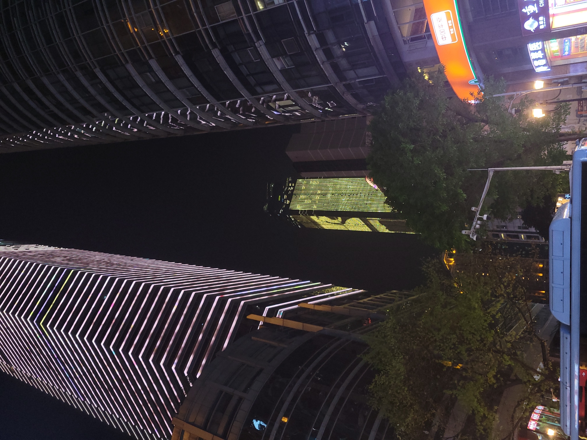 At night in Yuzhong District, China, two high-rise buildings are visible. The building on the left is illuminated with vertical lines of white light in a zig-zag pattern. The building on the right features rows of windows. A dark expanse of space separates the two structures, with a reflection of light visible within. Trees and a bus stop are in the right of the image. Signs with Chinese characters are visible.