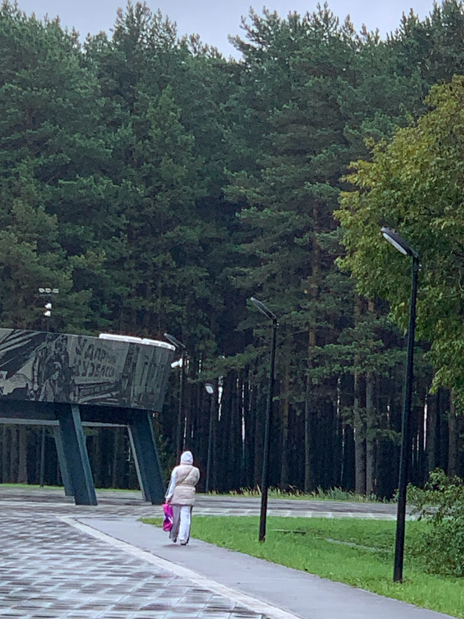 A lone person walks along a paved path, away from a large, dark, angular monument. The monument has a mural or inscription on its surface, with some text in Cyrillic. The person is wearing a light-colored puffer vest over a light-colored hooded sweatshirt and pants, and is carrying a pink and purple bag. They are walking on a paved walkway that leads away from the monument, bordered by a grassy area. In the background, a dense forest of tall pine trees dominates the scene. Several modern black lampposts are spaced along the path. The sky is overcast and the lighting suggests it is either early morning or late evening. The location is identified as the memorial complex "Krasnaya Gorka" in Prokopyevsk, Russia.