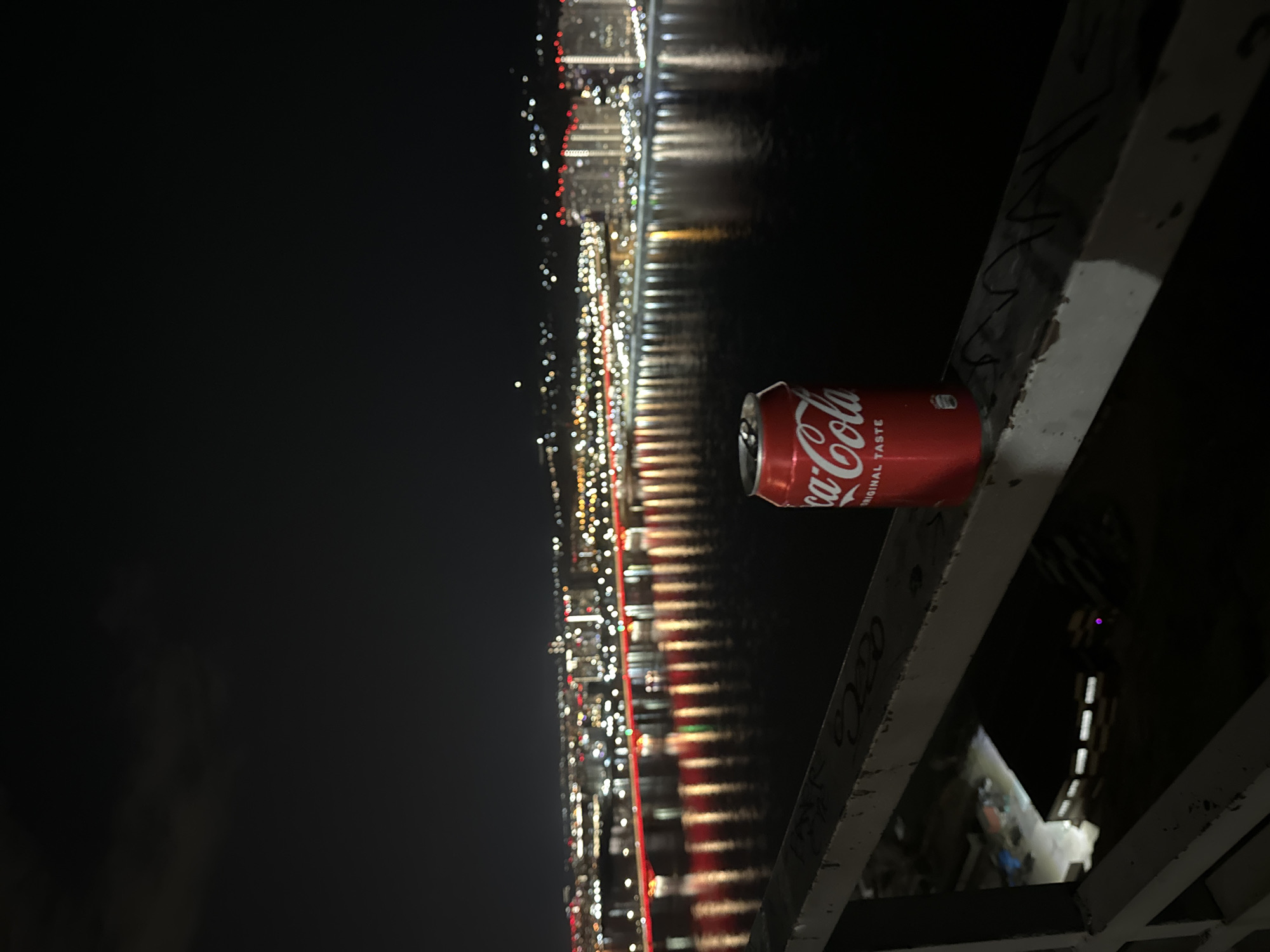 A horizontally oriented nighttime image shows a red can of Coca-Cola sitting on a white metal railing, with the reflection of city lights in the water visible in the background. The can is positioned on the right side of the frame. The lights are arranged in a linear fashion, suggesting the reflection of a city skyline. The focus is on the Coca-Cola can and the nearby railing. The scene appears to be set on a bridge overlooking a body of water. The presence of graffiti or markings on the railing indicates a public space. The location is the Kommunalny Bridge in Krasnoyarsk, Russia.