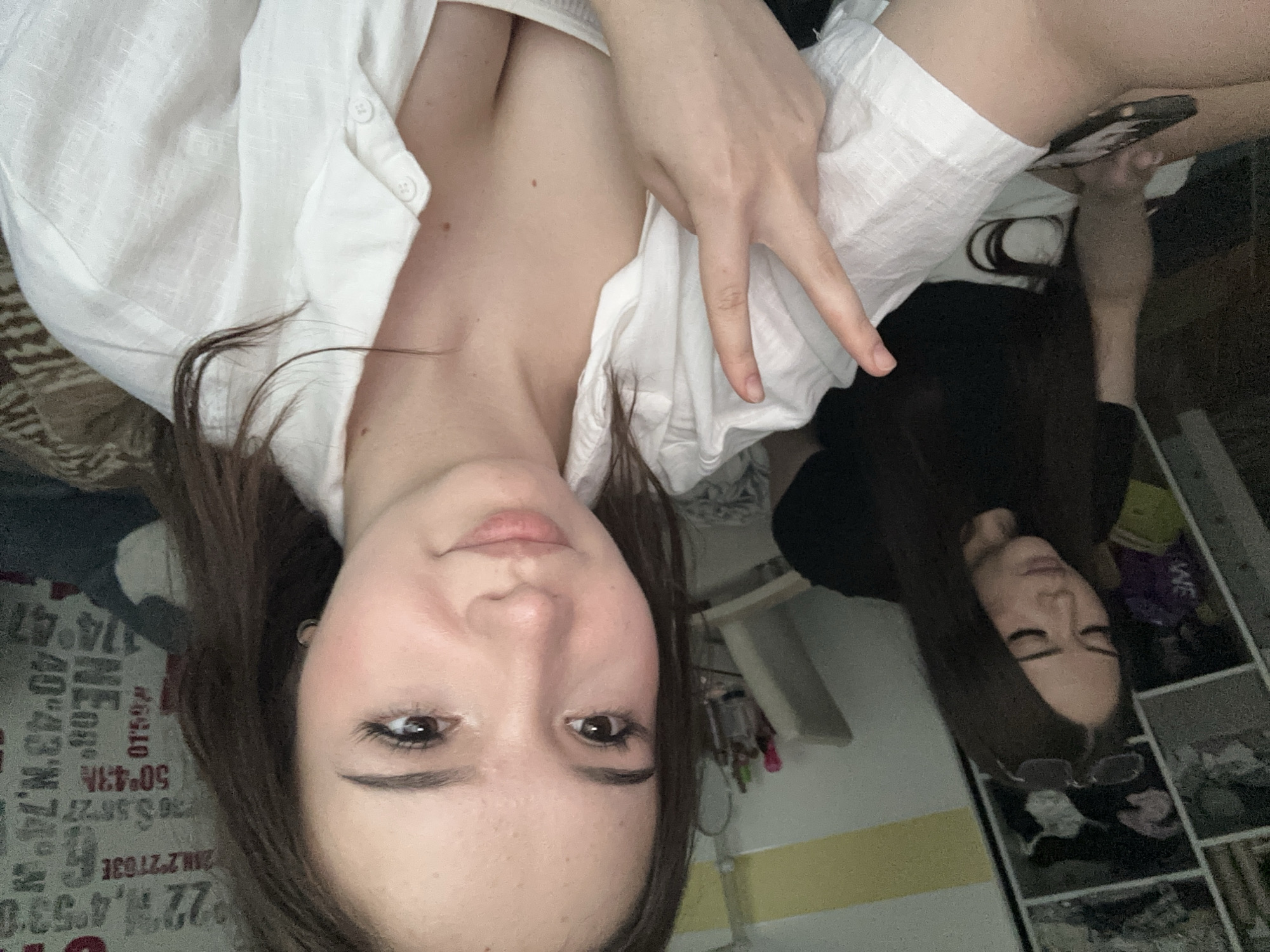 Two young women are indoors. The woman in the foreground, facing the camera, is wearing a white open-collared shirt over a white top. She has brown hair and is making a peace sign with her right hand. The woman in the background is wearing a black top and white shorts, and is looking down at her phone. She also has brown hair. They appear to be in a bedroom or living area, with a closet filled with clothes to the left and a wall decorated with map-like graphics in the background. The overall lighting suggests it is daytime. The specific location in Naberezhnye Chelny, Russia cannot be determined from the image.
