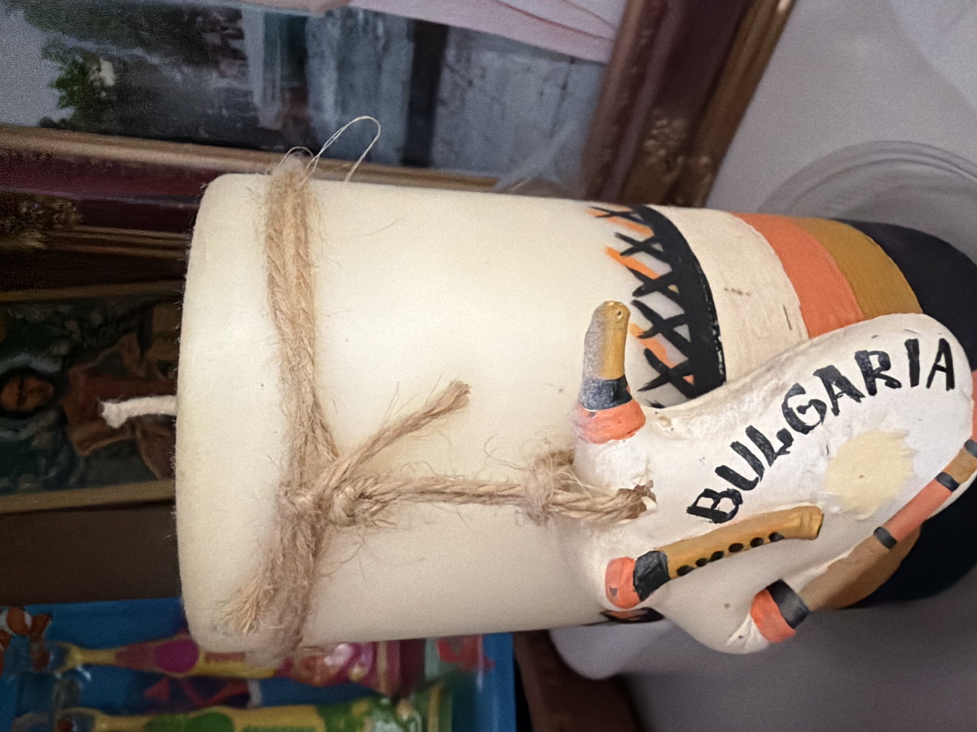 This is a close-up, eye-level shot of a white, cylindrical candle. The candle is adorned with a twine wrap around its upper section, tied in a knot with loose ends. A decorative tag, shaped like a stylized figure or instrument, hangs from the twine. This tag is painted with various colors, including orange, black, and gold, and bears the word "BULGARIA" in black lettering. The lower part of the candle itself is decorated with horizontal bands of orange, yellow, and black. In the background, blurred elements suggest a shelf or surface with other objects, including what appears to be a framed picture or painting. There is no discernible information within the image to indicate the specific location in the city of Pazardzhik, Bulgaria.