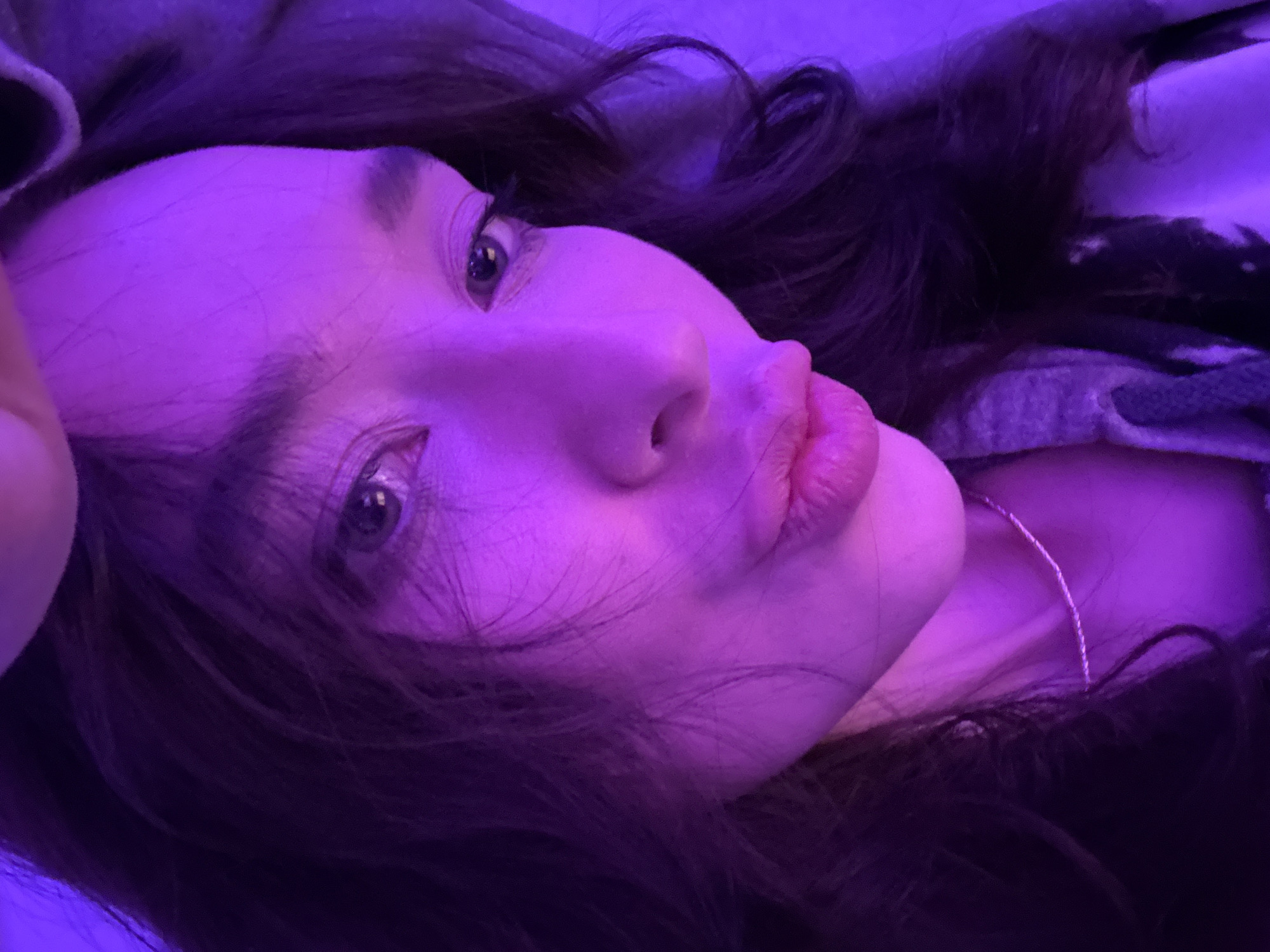 This close-up portrait features a person, likely female, with dark, wavy hair, illuminated by prominent purple light. The subject is shown from a slightly elevated and angled perspective, appearing to be lying down or reclining. Their face is turned upwards and slightly to the left, with the eyes looking in the same direction.
The purple lighting casts a strong, uniform hue over the entire scene, affecting skin tones and creating a moody or artistic ambiance. The person's lips are slightly parted, exhibiting a somewhat pouty expression. One eye is clearly visible, reflecting the purple light, with distinct eyelashes. Dark, flowing hair frames the face and spreads out around the head. A thin, silver-colored necklace or chain is visible resting on the person's neck.
On the upper right and left side, a textured, dark-patterned fabric, possibly a blanket or part of clothing like a hoodie, can be seen, contributing to the cozy or relaxed feel of the setting. The artificial lighting suggests an indoor scene, likely during evening or nighttime. There is no visible text in the image. The image is described as being from Moscow, Russia.