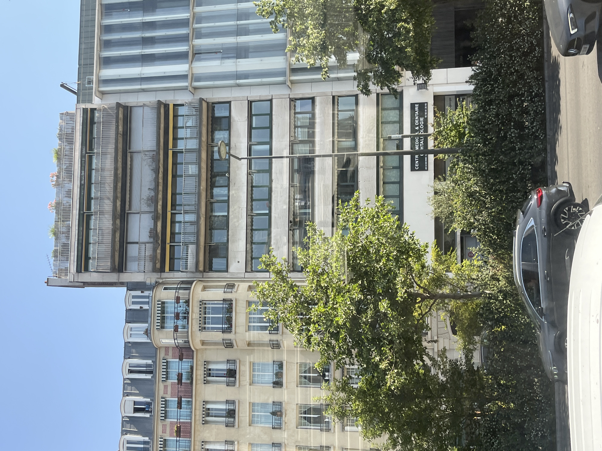 The image captures a street view in Neuilly-sur-Seine, France, on a clear, sunny day. The foreground features a street with two cars, one dark gray SUV on the left and a portion of a silver car on the right. Beyond the street, a row of buildings dominates the scene. The building on the left is a light-colored apartment building with traditional Parisian architecture, featuring mansard roofs, dormer windows, and balconies with black railings. Adjacent to it, to the right, is a modern, taller building with a concrete facade and numerous horizontal windows, some with closed blinds. Further to the right is a building with a striking facade composed of vertical, translucent panels. Lush green trees with dense foliage partially obscure the lower portions of the buildings. A streetlamp stands prominently in front of the concrete-faced building. The upper floors of the buildings show balconies and rooftop structures, one with greenery and what appear to be flowering plants. Signage on the ground floor of the concrete building indicates "CENTRE MEDICAL & DENTAIRE" and "HTALILOGIE," suggesting it houses a medical and dental center.