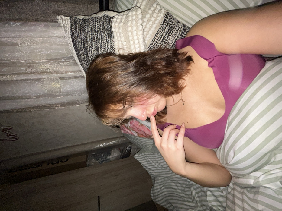 A person lies in bed, wearing a low-cut pink top with a cross necklace. Their head rests on a striped pillow next to a textured cushion. Their left hand is raised to their face, with a finger near their mouth. Striped bedding covers part of their body. The bed frame and the items beneath it are visible in the lower left corner. Another arm is visible to the top right of the image.