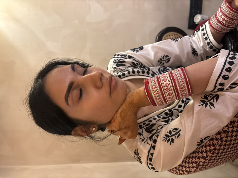 A single adult female is depicted indoors in the city of Howrah, India. She is seated, with her eyes closed and her head resting on her right hand. Her hair is dark and pulled back. She wears a white top featuring black floral patterns. Her right forearm is adorned with numerous red and white bangles, some with red floral designs and others embellished with small beads. Henna is visible on her right hand, and a ring is on her right ring finger. A light-colored stud earring is present on her left earlobe. The subject sits on a chair with a brown and white patterned fabric. The background is a plain, light-colored wall. A dark chair back and a partial white electrical outlet are visible to the right.