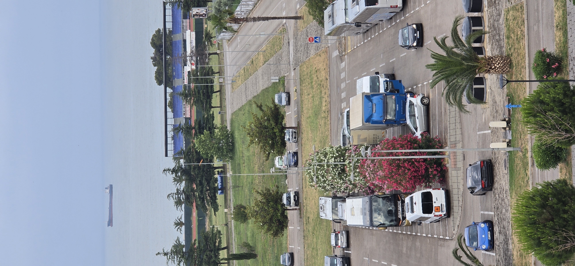 The image provides an elevated view of a coastal area in Bar, Montenegro, under an overcast or hazy daytime sky. In the foreground and extending into the midground, a wide road features multiple lanes and numerous parking areas. A variety of vehicles are present, including cars of different models and colors, several white vans, a blue Iveco truck, and a large white, gold, and black tour bus with "EURO SUNTC" clearly visible on its side. Some vehicles are in motion, while many others are parked.
The road is flanked by extensive green areas, featuring mature trees like palm trees and conifers, as well as vibrant flowering bushes in pink and white. Notable urban details include streetlights, bollards, a blue fire hydrant, a red "STOP" sign, and a pedestrian crossing sign.
Further into the midground, beyond the green spaces, rows of blue stadium seating are visible, suggesting the presence of a sports complex or arena adjacent to the coast. The background is dominated by the calm, light blue expanse of the sea, stretching towards the horizon. A large, dark-hulled cargo ship with a lighter superstructure can be seen in the distance on the water. The overall scene depicts typical daily activity in a Mediterranean port town.