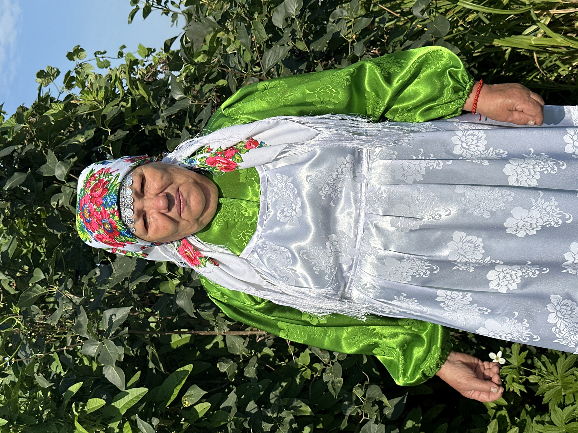 In an outdoor setting with lush green foliage and a clear blue sky, a woman stands facing the camera. She is wearing traditional Russian attire. Her head is adorned with a colorful floral headscarf, accented with silver trim. A white scarf with floral embroidery and fringe is draped around her shoulders. She is dressed in a bright green blouse with puffed sleeves and a white apron with a floral pattern. Her right hand is slightly raised, holding a small white flower. A red string bracelet is visible on her right wrist. The background consists of dense green bushes and trees. The woman appears to be in the village of Sredneye Kechevo, Russia.