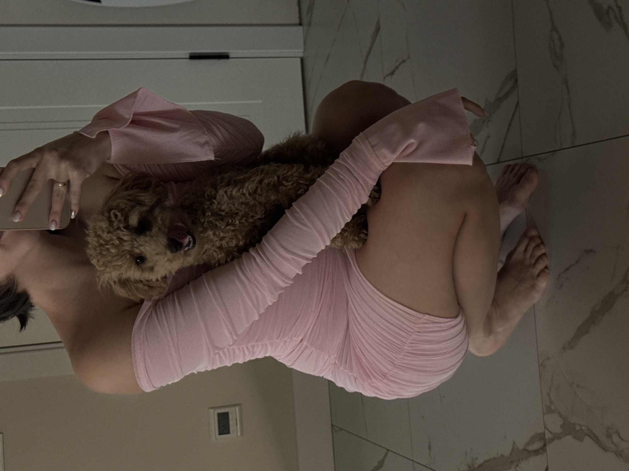 An indoor mirror selfie captures a person in a light pink ruched mini dress with long sleeves, holding a small, curly-haired, light brown dog. The person's left hand, adorned with a gold ring and neatly manicured nails, holds a light-colored smartphone, indicating they are taking the photo. Their bare feet are visible on a light-colored, marble-patterned tiled floor. The dog is positioned on the person's upper body, with its mouth slightly open and tongue visible, giving it an expressive, possibly panting, look. The background behind the person features a white door or wall paneling. The overall lighting is dim, suggesting an indoor setting, possibly in the evening or with low natural light. A dark rectangular object, possibly an outlet or switch, is visible on the wall. The scene suggests a casual moment shared between a pet owner and their dog within a residence in Pervomaysky, Russia. No visible text is present in the image.