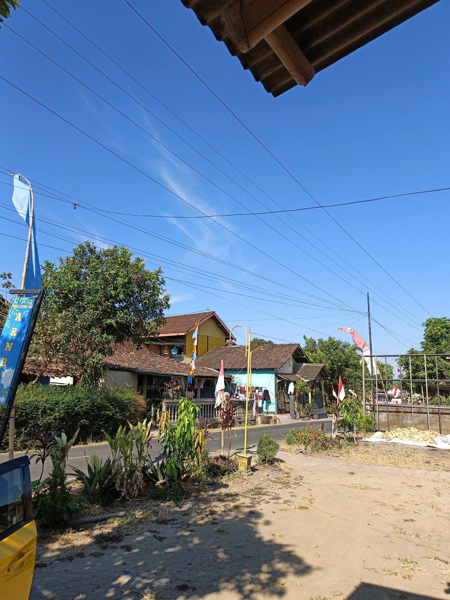 The image depicts a roadside scene in Jatinom, Indonesia, on a clear, sunny day. The foreground is dominated by a dirt and gravel area with scattered vegetation and shadows cast by an unseen structure, possibly a building's overhang. A bright yellow car door is visible on the left side of the frame, partially obscuring a blue banner with white lettering that reads "WELCOME TO ARNB".
Beyond the foreground, a paved road runs horizontally across the image. Along the roadside, there are various plants and small trees. Several houses with terracotta tiled roofs are visible, one painted teal and another a pale yellow. Flags, including the Indonesian flag (red and white), are displayed in front of some of the houses and on poles.
Electrical wires crisscross the sky, which is a vibrant blue with a few wispy clouds. A large, leafy green tree stands prominently between two of the houses. In the distance, a metal fence encloses an area where a large pile of light-colored, possibly dried, goods is covered by a white tarp. The overall impression is that of a rural or semi-urban residential area. No people are visible in the image.