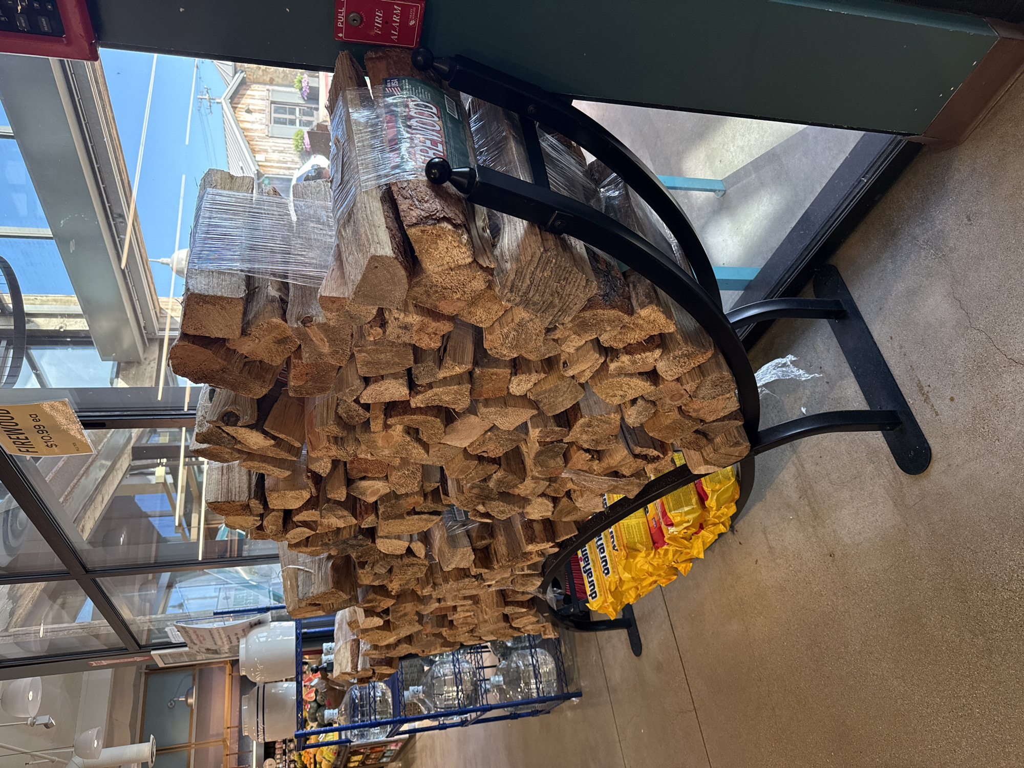 The image shows a large stack of firewood on display in what appears to be an outdoor or semi-outdoor retail space, possibly a grocery store or garden center, located in Malibu, United States of America. The firewood is neatly stacked and wrapped in clear plastic. Some bundles of Duraflame firestarters are visible at the base of the firewood display. To the left, there are shelves with large plastic jugs of water. In the background, through a large window, a rustic building with a wooden exterior is visible under a clear blue sky. A red fire alarm box is mounted on a dark green pillar to the right of the image. The flooring is a concrete surface. There are no people visible in the scene.