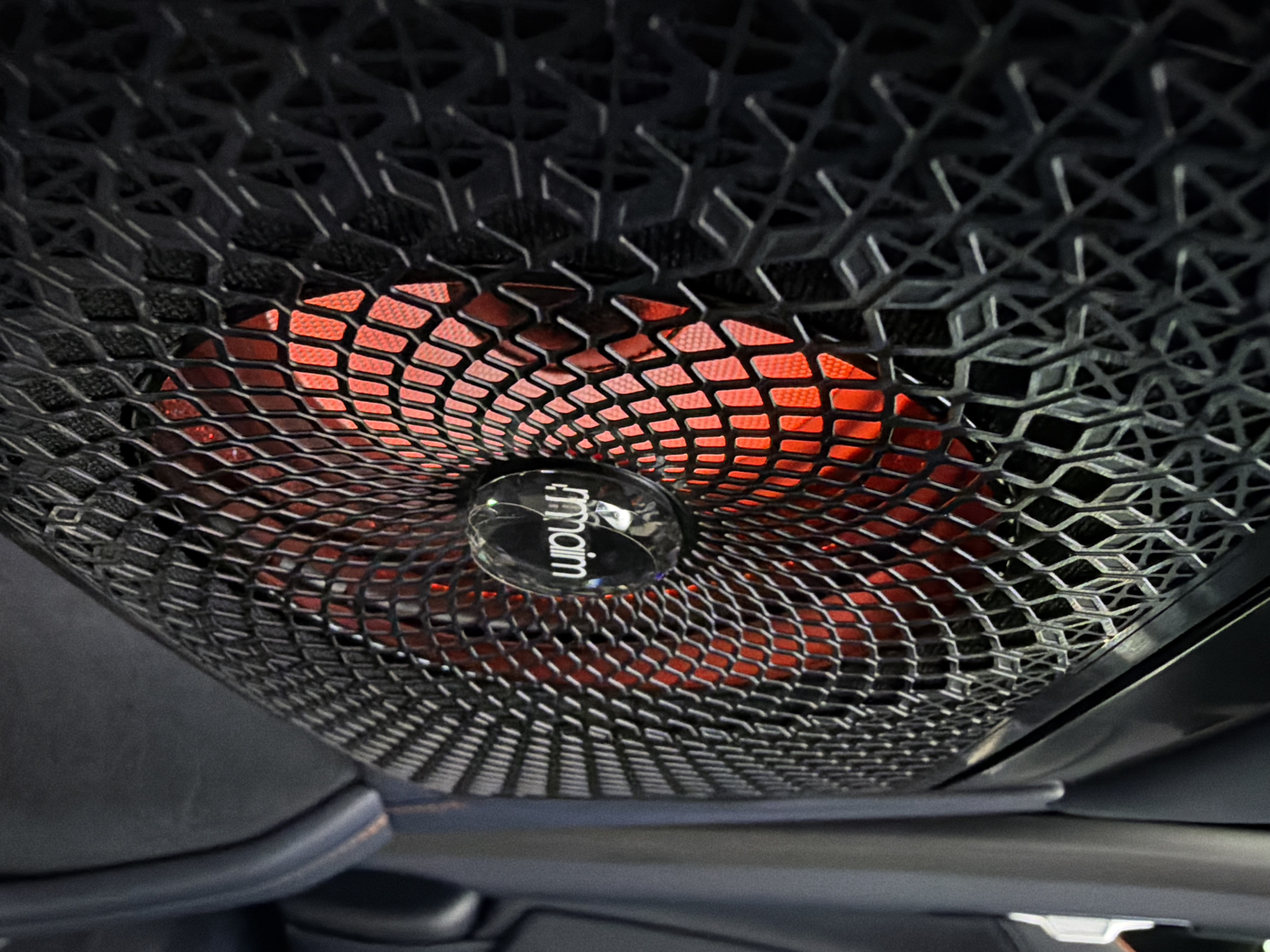 The image shows a close-up of a Naim speaker grille, likely inside a vehicle. The grille is black with a diamond-shaped pattern. Behind the grille, a red/orange light is visible, illuminating the speaker cone area. The Naim logo is visible on the center of the speaker. The surrounding environment is blurred, suggesting the speaker is located in a car's interior. There are no people visible. The location within Krasnoyarsk, Russia cannot be determined from the image.
