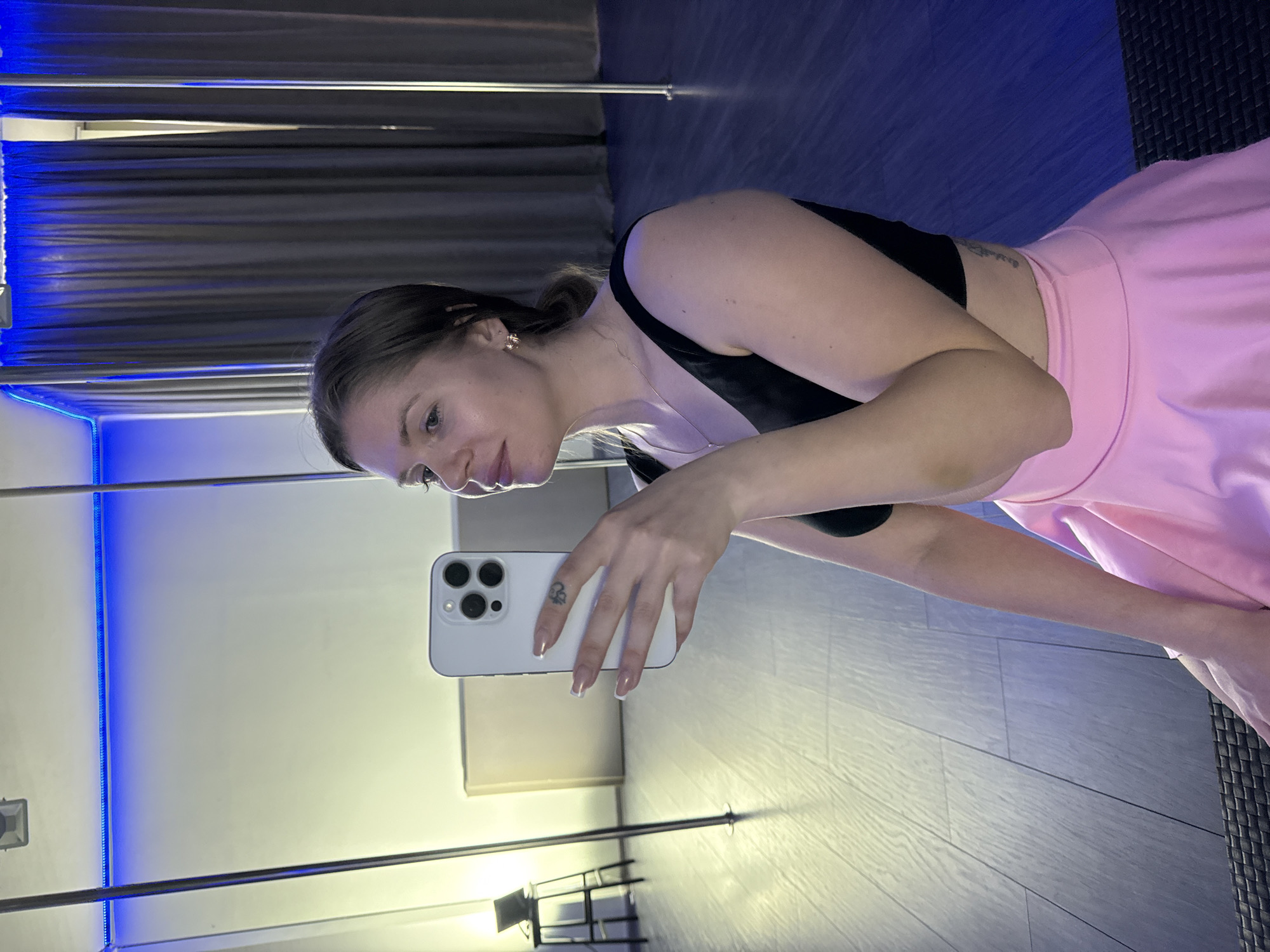 A woman is taking a selfie in what appears to be a dance studio. She is wearing a black top and a pink skirt. The background includes mirrors with blue LED lighting along the edges, dark curtains, and what appears to be a floor with some tiled or patterned areas. The location is likely a dance studio, potentially in Batumi, Georgia. The focus of the image is on the woman and her surroundings in the studio setting.