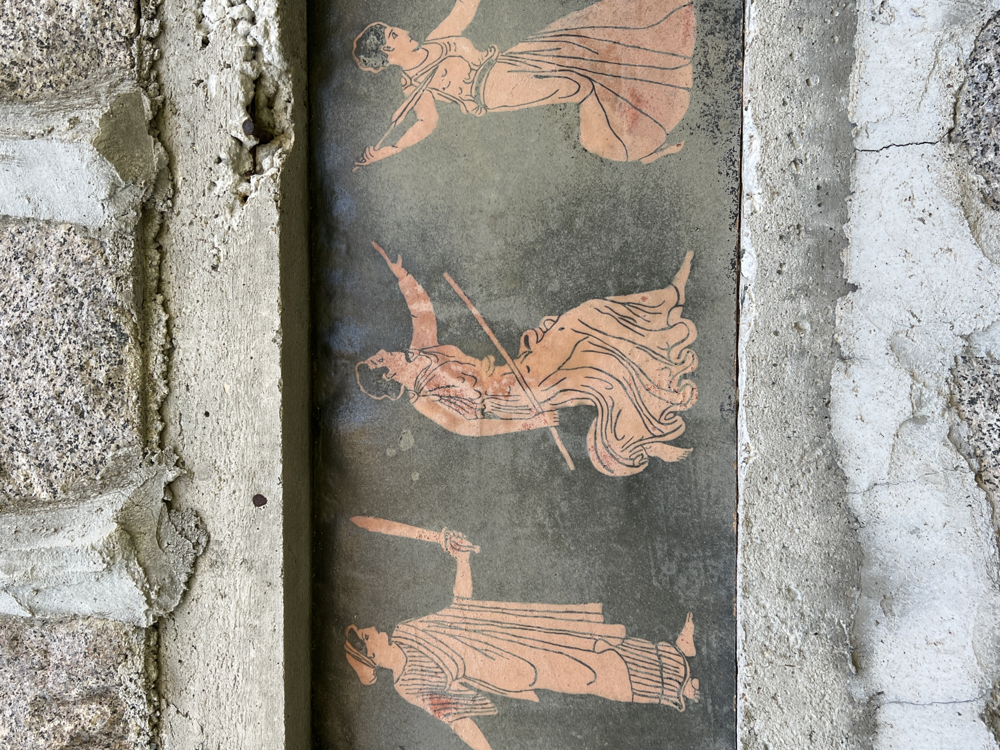 The image shows a bas-relief carving on a textured, dark surface. Three figures in classical attire are depicted in profile. The leftmost figure, facing right, holds a sword aloft in its right hand. The middle figure, also facing right, holds a staff in its right hand and gestures with its left. The rightmost figure, facing left, holds a staff with both hands, with its left arm extended upwards. The figures are rendered in an orange-pink hue with black outlines. The carving is set within a concrete structure, with rough, light-colored stone visible above and below the carved panel. The concrete is weathered and shows signs of age and repair. The overall impression is of an ancient or historical art piece integrated into a rustic architectural element, possibly located outdoors in a place like Sinemorets, Bulgaria, given the natural stone and weathered concrete.