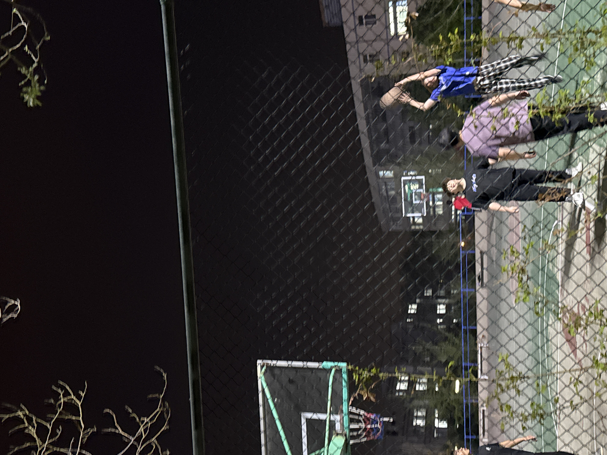 This image shows a basketball court in Zhongtun, China, viewed through a chain-link fence. Three people are on the court; one is wearing a blue shirt and patterned pants, another is wearing a purple shirt, and a third is wearing a black shirt with a red detail. A basketball hoop is visible. The court is illuminated with buildings visible in the background. The background contains a dark sky. There are trees along the left side of the fence.
