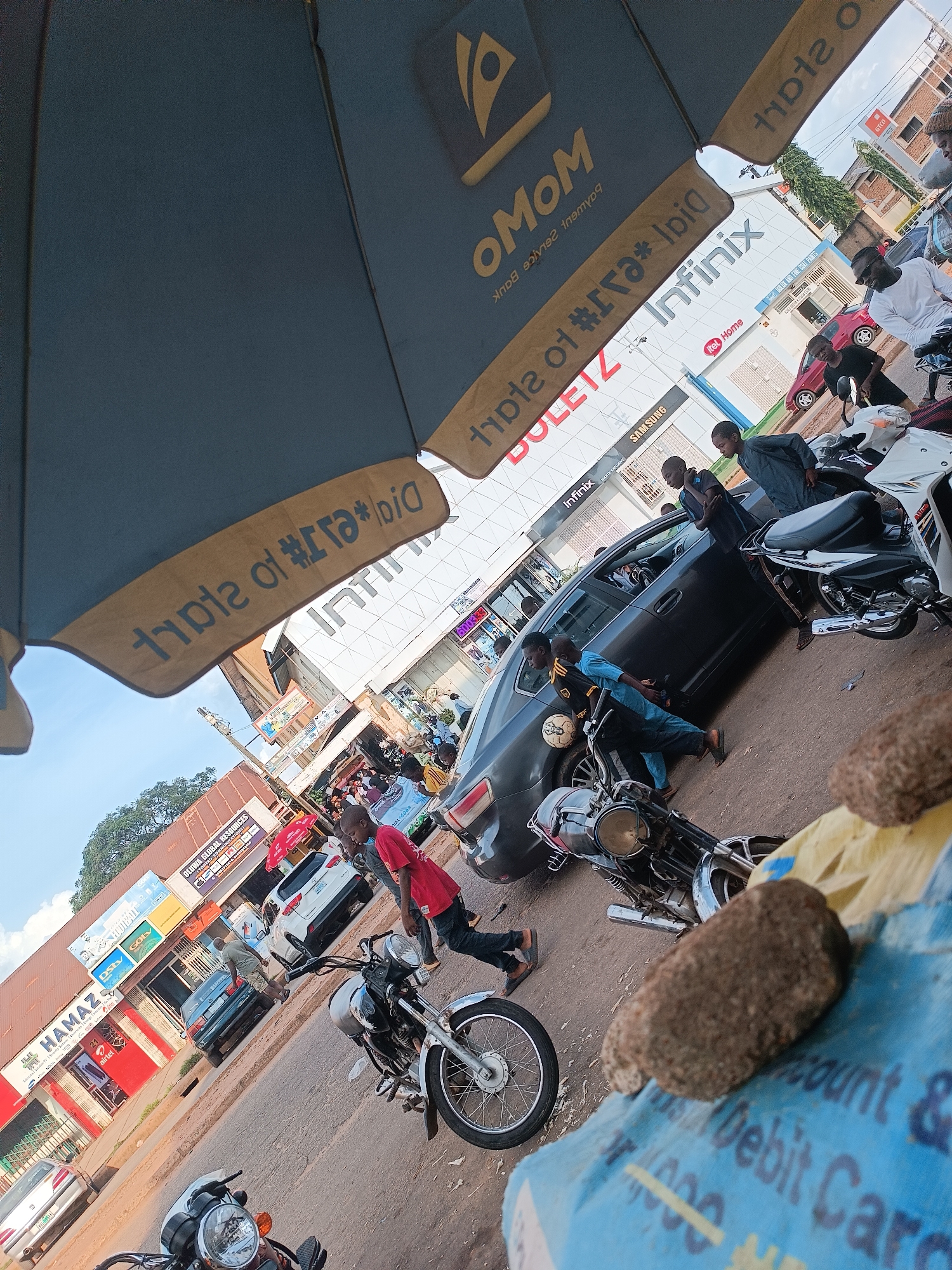 The image captures a bustling street scene in Jos, Nigeria, likely near a commercial area given the presence of shops and advertisements. An open blue umbrella dominates the foreground, partially obscuring the view of the sky and the surrounding buildings. Visible on the umbrella are yellow text and logos.
In the mid-ground, a dark gray sedan is parked on the street, with several people around it. One individual in a blue shirt and black shorts is holding a soccer ball. Another person in a dark outfit is standing near the car's driver's side. Further down the street, a white car and a dark green car are also visible, along with more people and motorcycles.
Shops line the street, with signs indicating businesses such as "HAMAZ," "DSTV," "SONY," and "OLUWA GLOBAL RESOURCES." An advertisement for "Infinix" is prominently displayed on a building. Several motorcycles are parked or in motion on the street, indicating a common mode of transportation.
The overall environment suggests a vibrant, active urban setting with commercial activity and daily life unfolding. The image is shot from a slightly upward angle, contributing to a dynamic perspective of the scene.