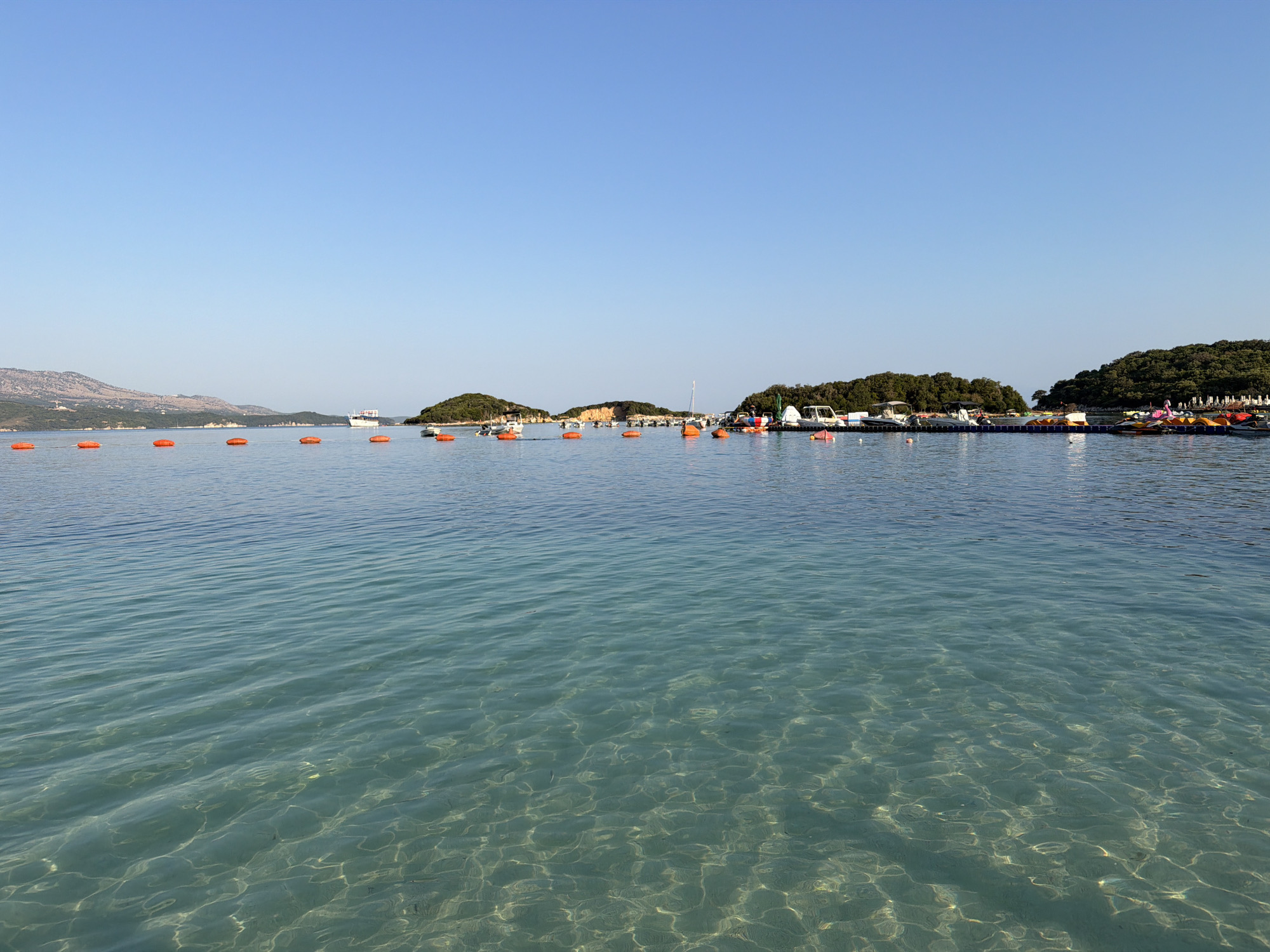 This image shows a bright, sunny day at a beach in Ksamil, Albania. The foreground is dominated by clear, shallow turquoise water, with sunlight reflecting off the sandy seabed. A line of orange buoys stretches across the water, marking a boundary. In the mid-ground, several small islands covered in lush green trees dot the horizon. A few boats, including a white passenger ferry and several smaller motorboats, are moored in the water near the islands. Some of these boats appear to be used for rentals, with brightly colored pedal boats visible. The background consists of a clear, light blue sky with no clouds. The overall scene depicts a tranquil and idyllic coastal environment.