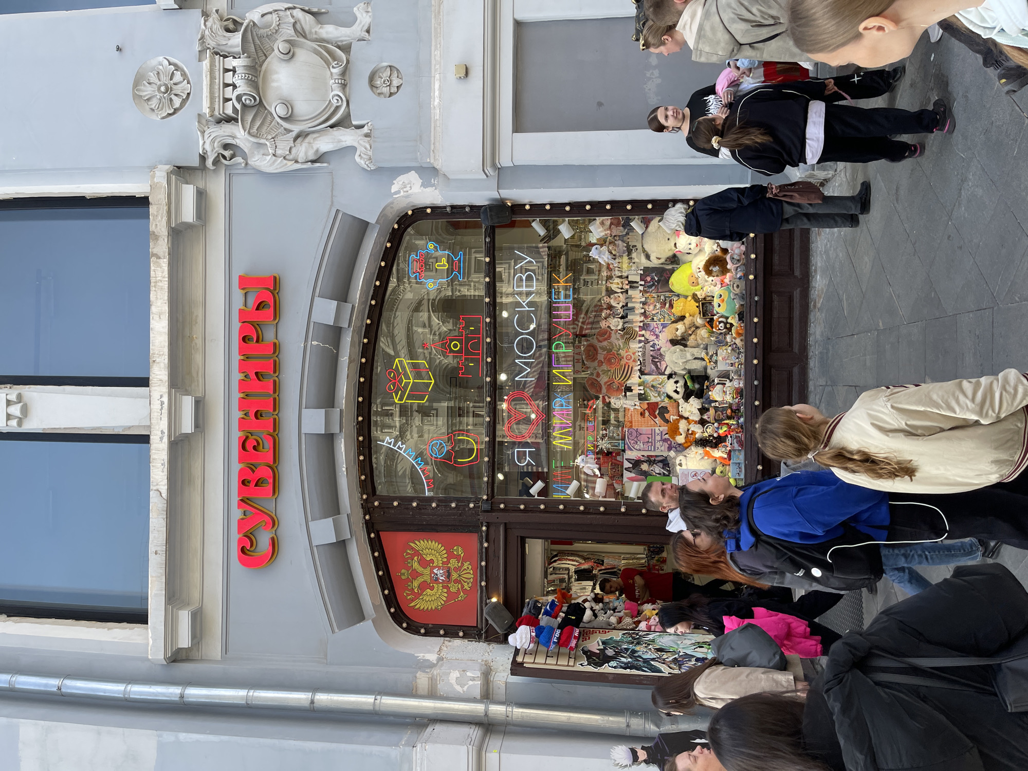 The image depicts the exterior of a shop in Moscow, Russia, called "Сувениры" (Souvenirs). The shop has a curved, arched storefront window filled with various plush toys and souvenirs. Neon signs in the window include depictions of Russian icons and the name of the shop. A Russian double-headed eagle emblem is displayed above the entrance. A crowd of people is gathered in front of the shop, looking at the display. They are dressed in everyday clothing, and appear to be tourists or shoppers. The building facade is decorated with ornate carvings.