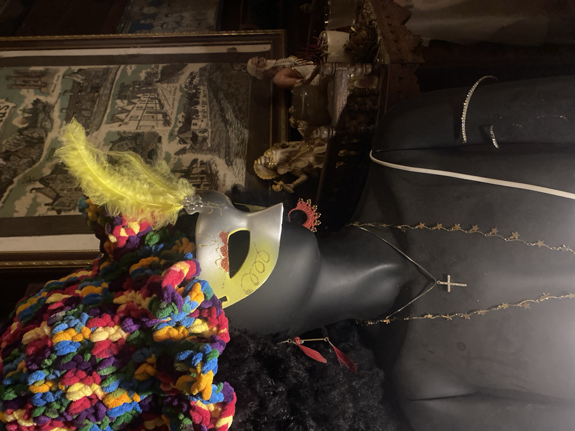 A close-up photograph features a black mannequin adorned with various accessories and decorations. A masquerade mask with a yellow feather rests atop a colorful, textured garment resembling yarn. A gold-colored necklace with a cross pendant and a second necklace decorated with star shapes are draped around the mannequin's neck. The mannequin also wears jewelry, including what appears to be a beaded bracelet. In the background, a framed image and other decorative objects are partially visible. The location is Baan Don Sai, Thailand.