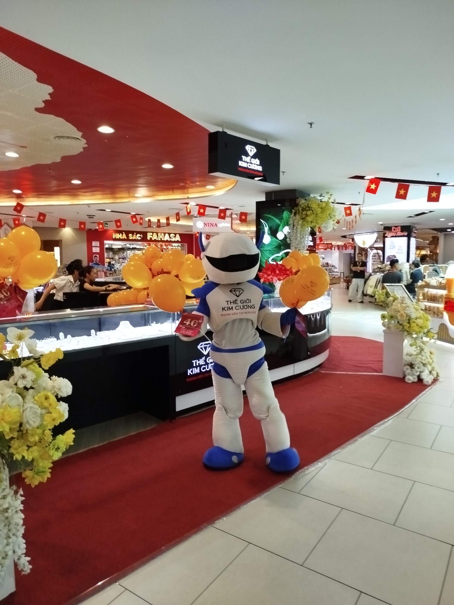 This vibrant image captures an indoor commercial space, likely a shopping mall in Da Lat, Vietnam, decorated for a special event or promotion.
The main subject is a white and blue robot-like mascot standing on a red carpet in front of a jewelry store. The mascot holds a bunch of orange balloons in its left hand and a promotional card in its right hand. The balloons also feature the store's branding.
The setting is a well-lit shopping area. The jewelry store, identified by the signage "THẾ GIỚI KIM CƯƠNG" (The World of Diamonds) and "THÀNH VIÊN TẬP ĐOÀN DOJI" (Member of DOJI Group), is prominent with its black display cases showcasing jewelry. Behind the counter, two female staff members are visible. Numerous orange balloons are arranged throughout the store's display. The store entrance features a red carpet flanked by white and yellow floral arrangements.
In the background, other retail spaces are visible, including "NHÀ SÁCH FAHASA" (FAHASA Bookstore) and a store with "NINA" signage. The ceiling and upper walls are festively decorated with strings of small red flags featuring a yellow star, characteristic of the Vietnamese national flag, reinforcing the location context. Part of the ceiling is a distinct red shape resembling a map.
The activity appears to be a promotional event or grand opening, indicated by the mascot, balloons, red carpet, and special decorations. The mascot holds a card displaying "40" and additional text, possibly indicating a discount or anniversary. The overall mood is lively and inviting. The scene is brightly lit, suggesting daytime inside the mall.