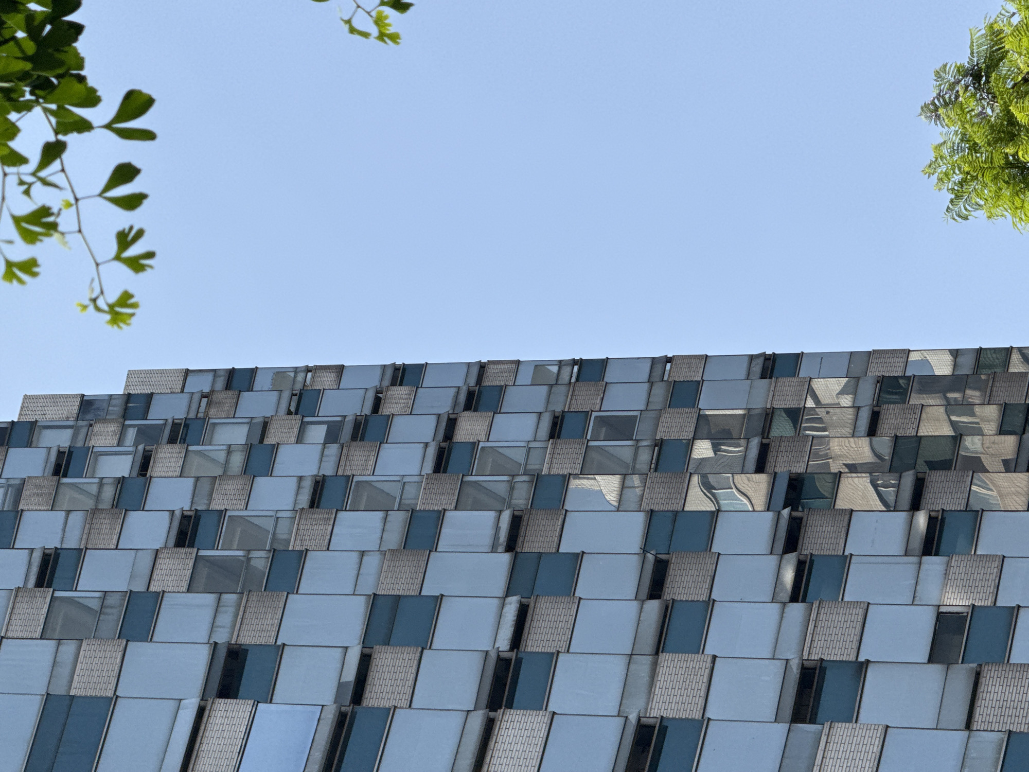 A low-angle shot of a modern building with a unique exterior of tiered, reflective panels, observed under a clear sky. Green leaves from trees frame the top edges of the image. The building's location is assumed to be within Jinjiang District, China. The dominant features are the reflective panels that create a step-like pattern on the building's facade. The panels are alternating in color from light blue to gray. The light reflecting off the panels indicates the weather is sunny.