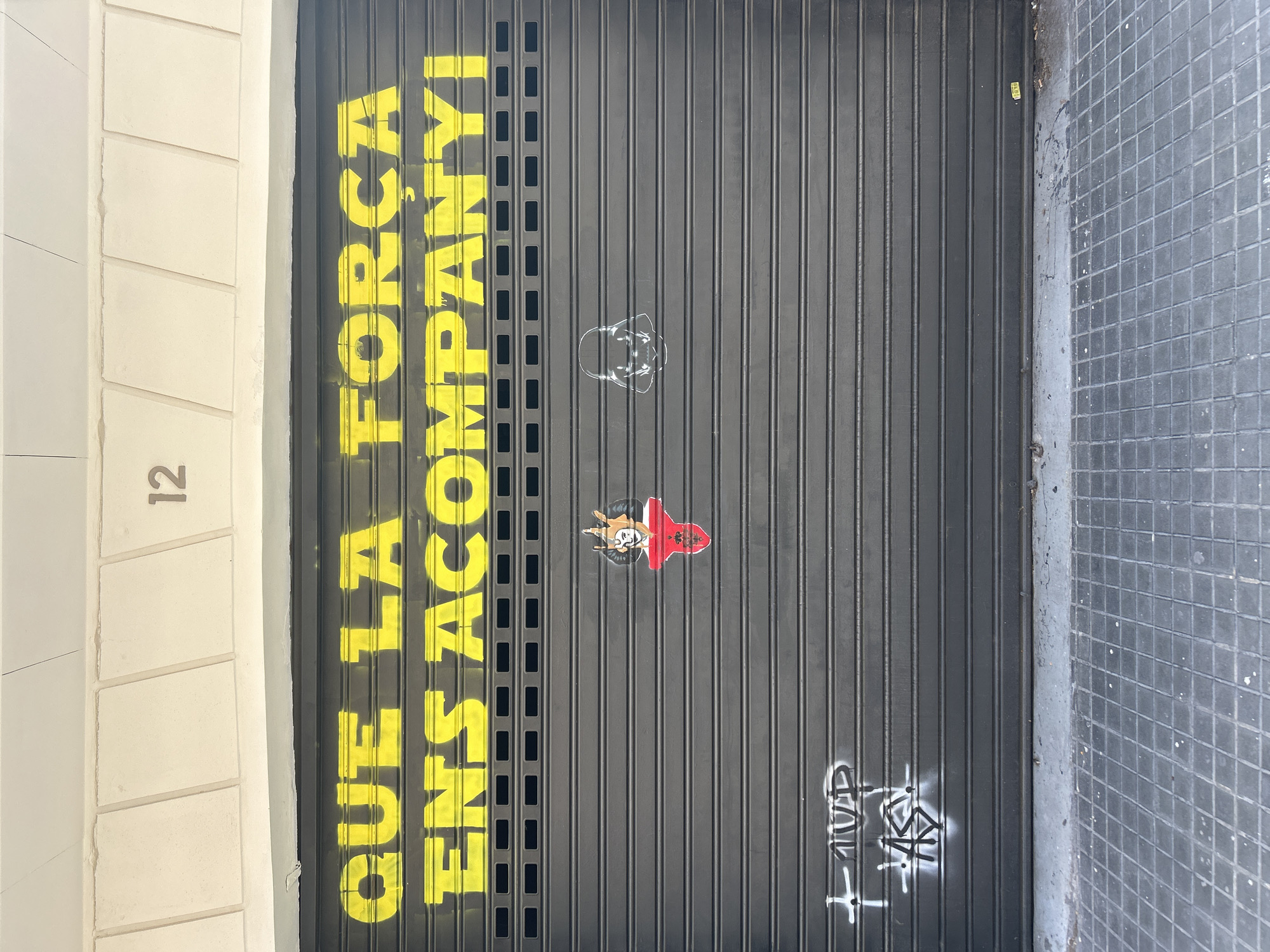 The image shows a closed gray corrugated metal garage door on a building. Above the garage door, the number "12" is visible on a cream-colored facade with rectangular stone accents. Yellow spray paint lettering on the garage door reads "QUE LA FORÇA ENS ACOMPANYI," which translates to "May the Force be with you" in Catalan. To the left of the text, a small sticker depicts a cartoonish devil-like figure with horns and a red collar. To the right of the text, a white stencil of Darth Vader's helmet is visible. In the lower left corner of the garage door, white graffiti includes a cross shape and abstract lettering. The ground in front of the garage door is paved with dark gray square tiles. The overall scene suggests an urban environment in Barcelona, Spain, with the graffiti and building details being characteristic of the city.